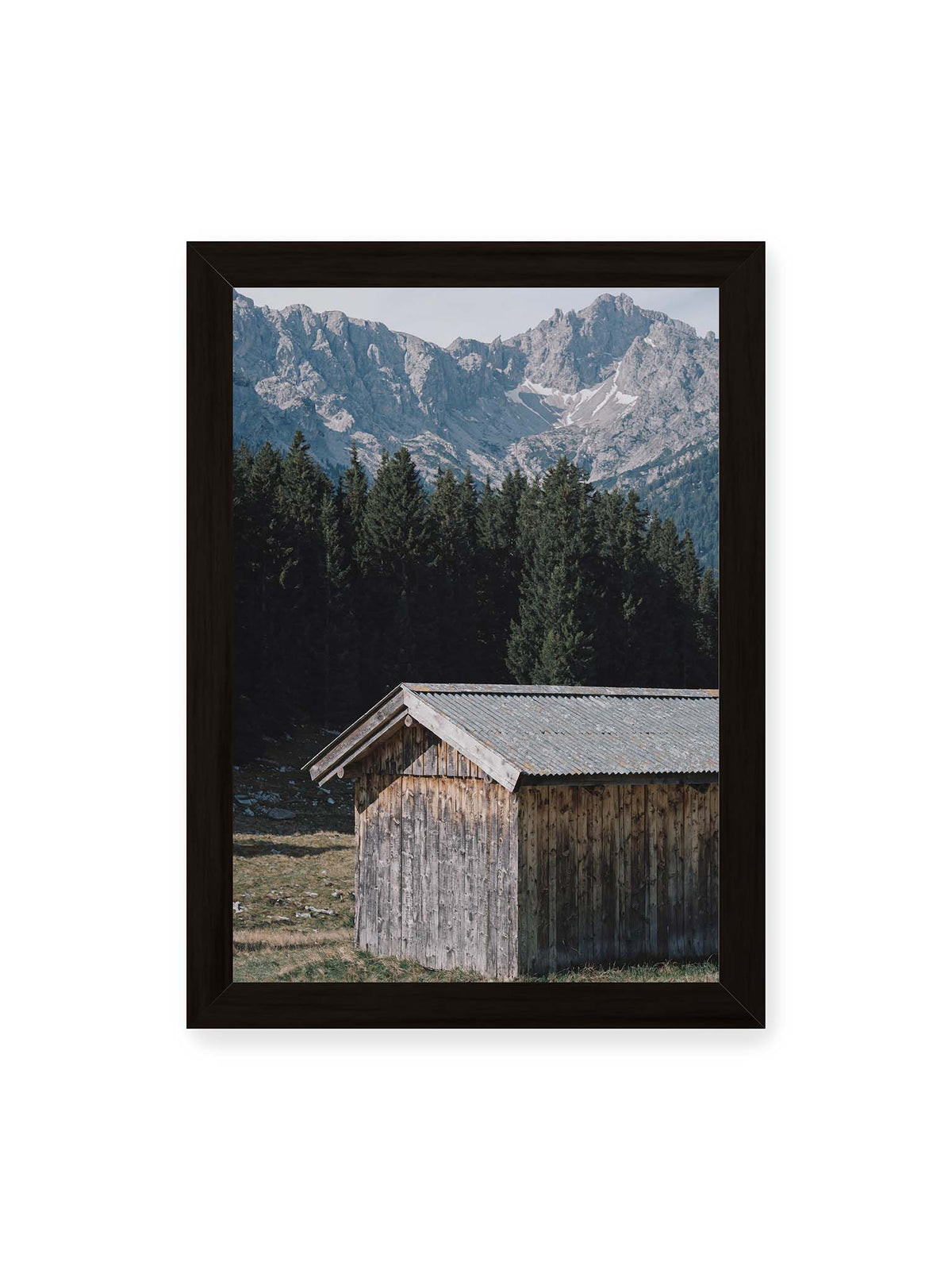 21x30 cm, schwarzer Rahmen – Verwitterte Holzhütte vor dunklen Nadelbäumen und einer hellen, zerklüfteten Bergkulisse.