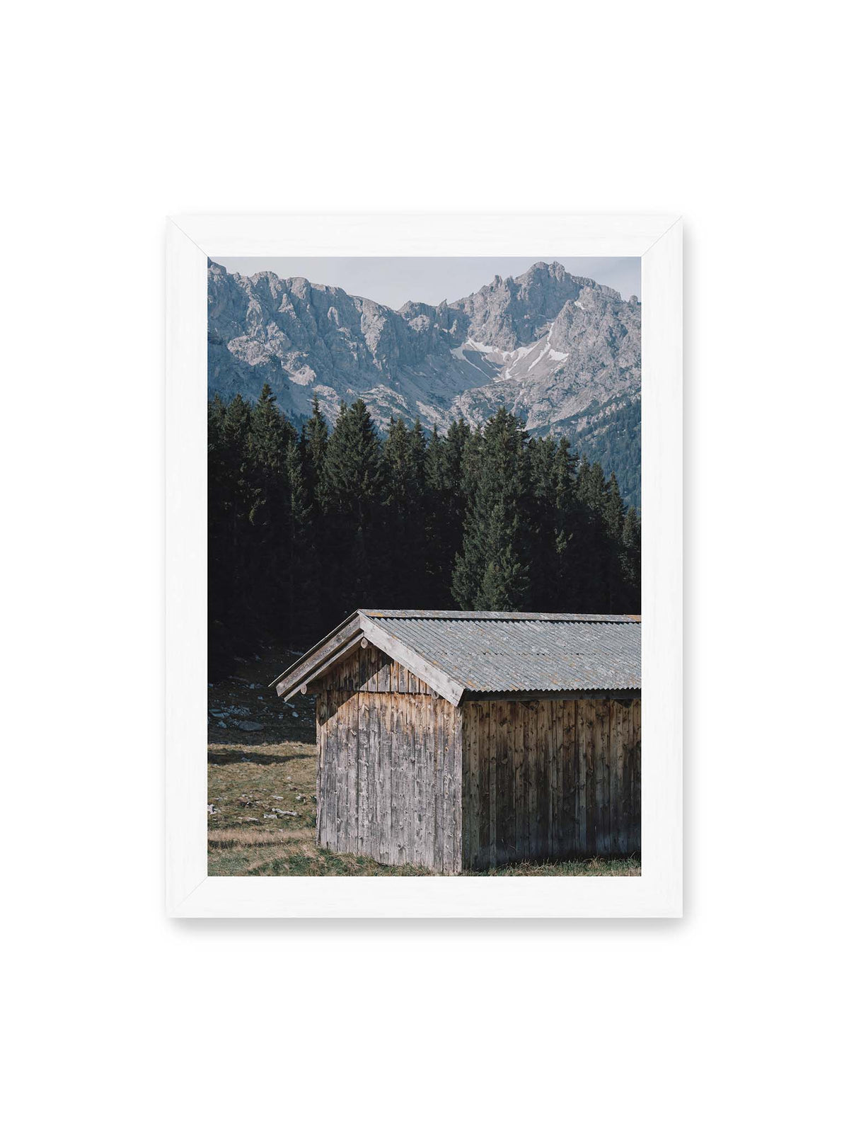 21x30 cm, weißer Rahmen – Verwitterte Holzhütte vor dunklen Nadelbäumen und einer hellen, zerklüfteten Bergkulisse.