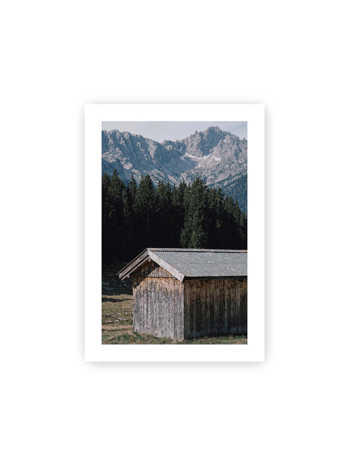 21x30 cm, weißer Galerie-Rand – Verwitterte Holzhütte vor dunklen Nadelbäumen und einer hellen, zerklüfteten Bergkulisse.
