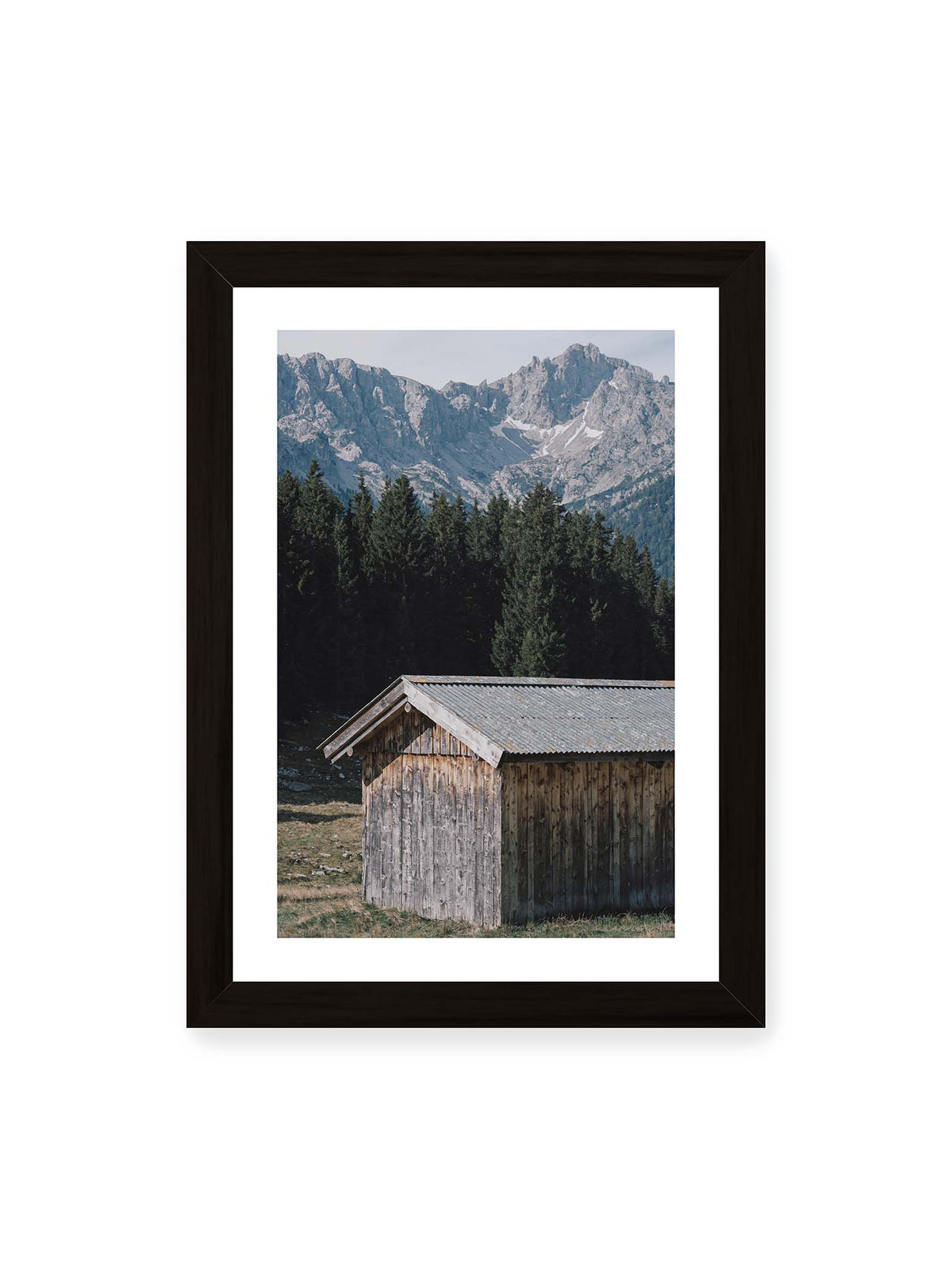 21x30 cm, weißer Galerie-Rand, schwarzer Rahmen – Verwitterte Holzhütte vor dunklen Nadelbäumen und einer hellen, zerklüfteten Bergkulisse.