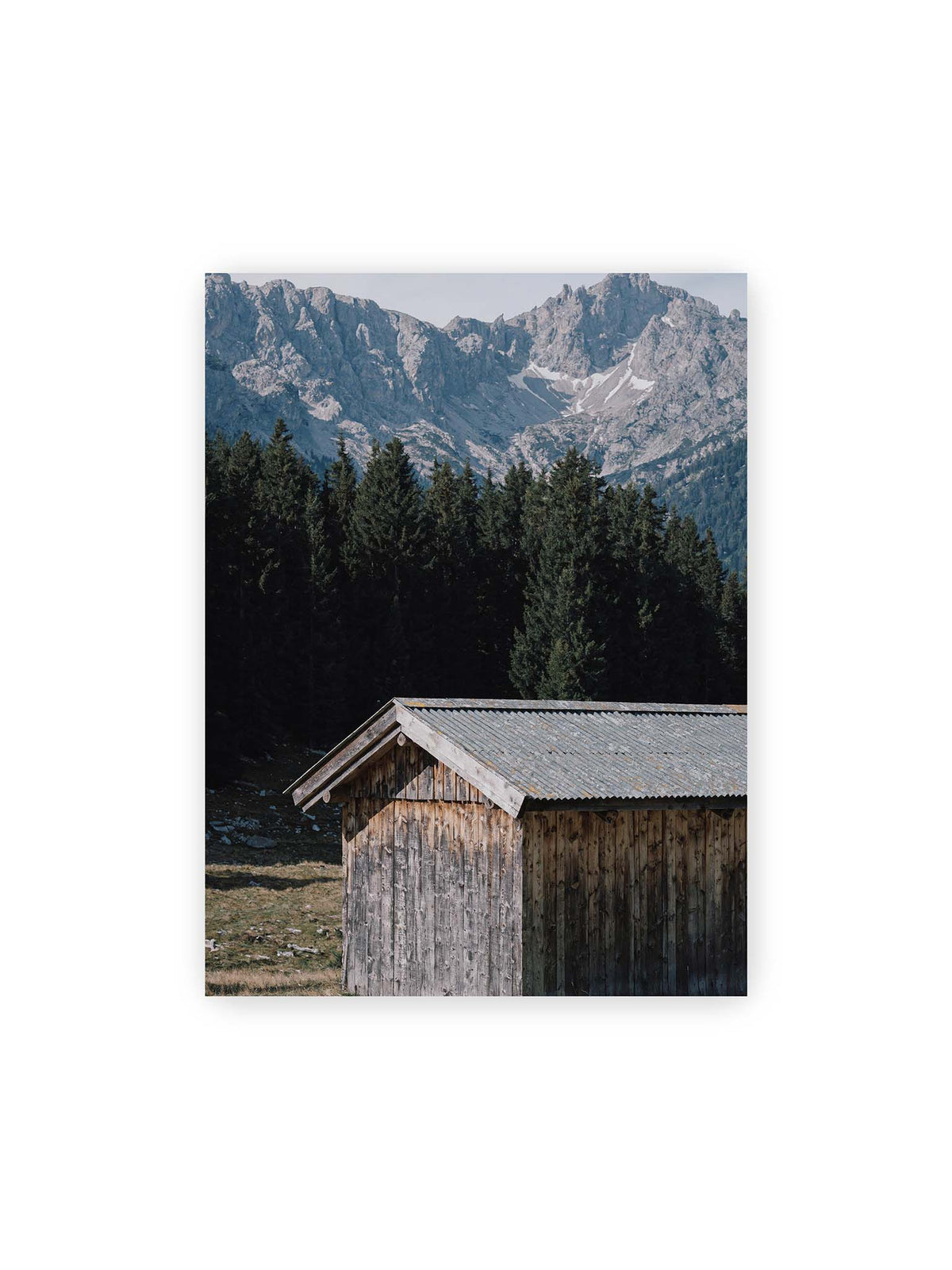 30x40 cm – Verwitterte Holzhütte vor dunklen Nadelbäumen und einer hellen, zerklüfteten Bergkulisse.
