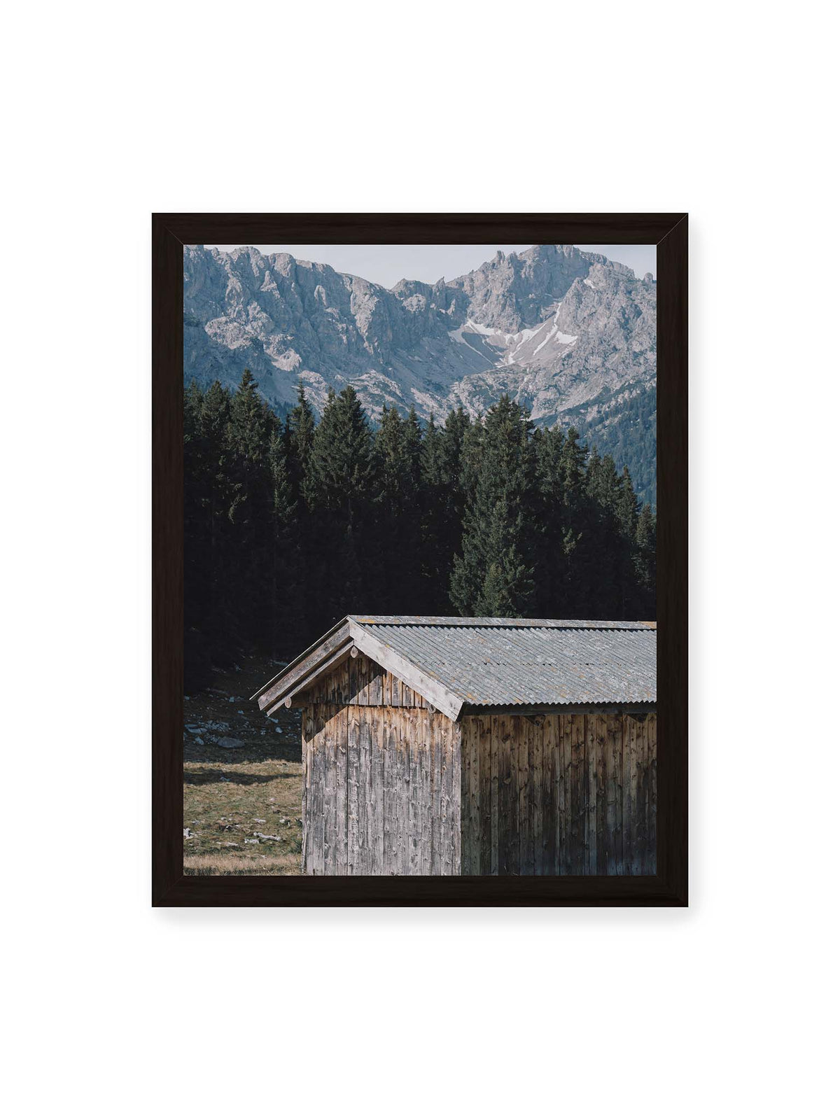 30x40 cm, schwarzer Rahmen – Verwitterte Holzhütte vor dunklen Nadelbäumen und einer hellen, zerklüfteten Bergkulisse.