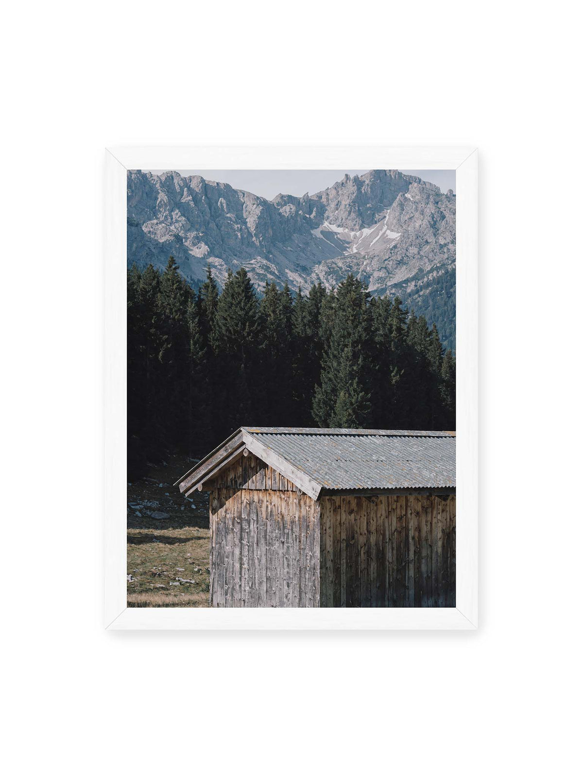30x40 cm, weißer Rahmen – Verwitterte Holzhütte vor dunklen Nadelbäumen und einer hellen, zerklüfteten Bergkulisse.
