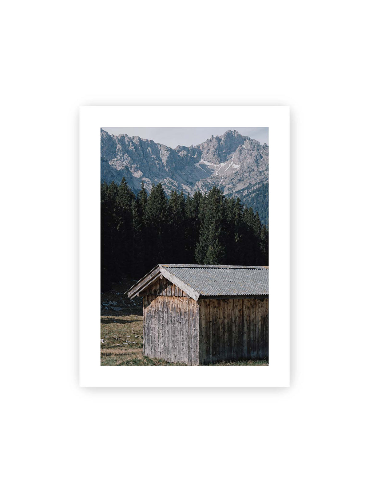 30x40 cm, weißer Galerie-Rand – Verwitterte Holzhütte vor dunklen Nadelbäumen und einer hellen, zerklüfteten Bergkulisse.