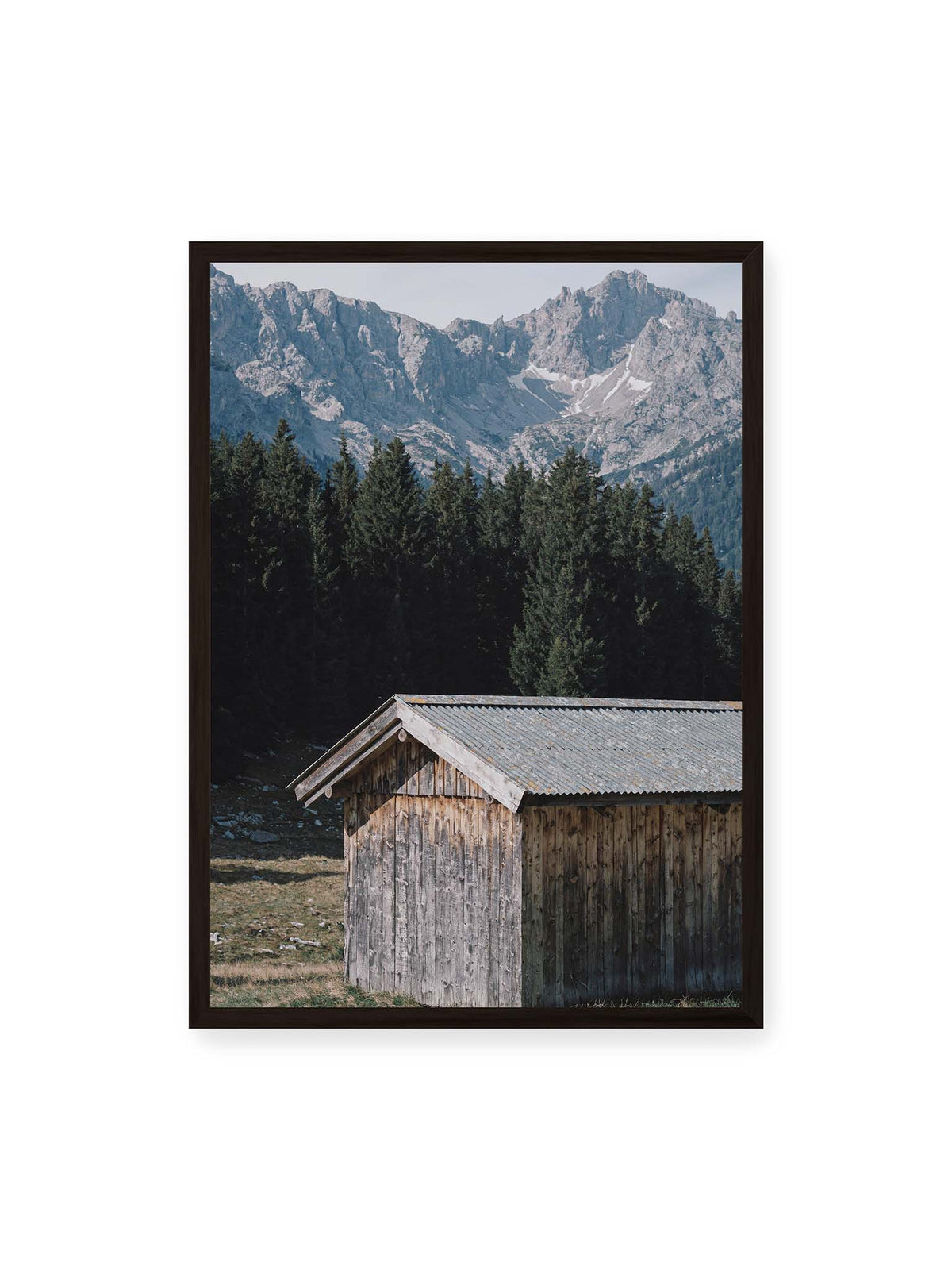 50x70 cm, schwarzer Rahmen – Verwitterte Holzhütte vor dunklen Nadelbäumen und einer hellen, zerklüfteten Bergkulisse.
