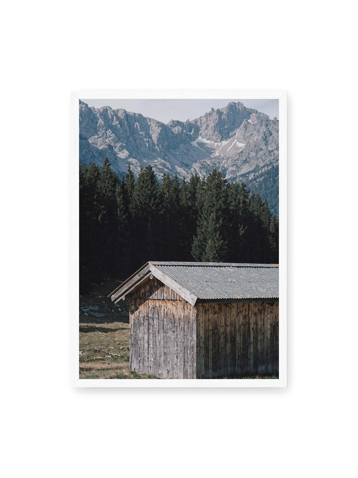 50x70 cm, weißer Rahmen – Verwitterte Holzhütte vor dunklen Nadelbäumen und einer hellen, zerklüfteten Bergkulisse.