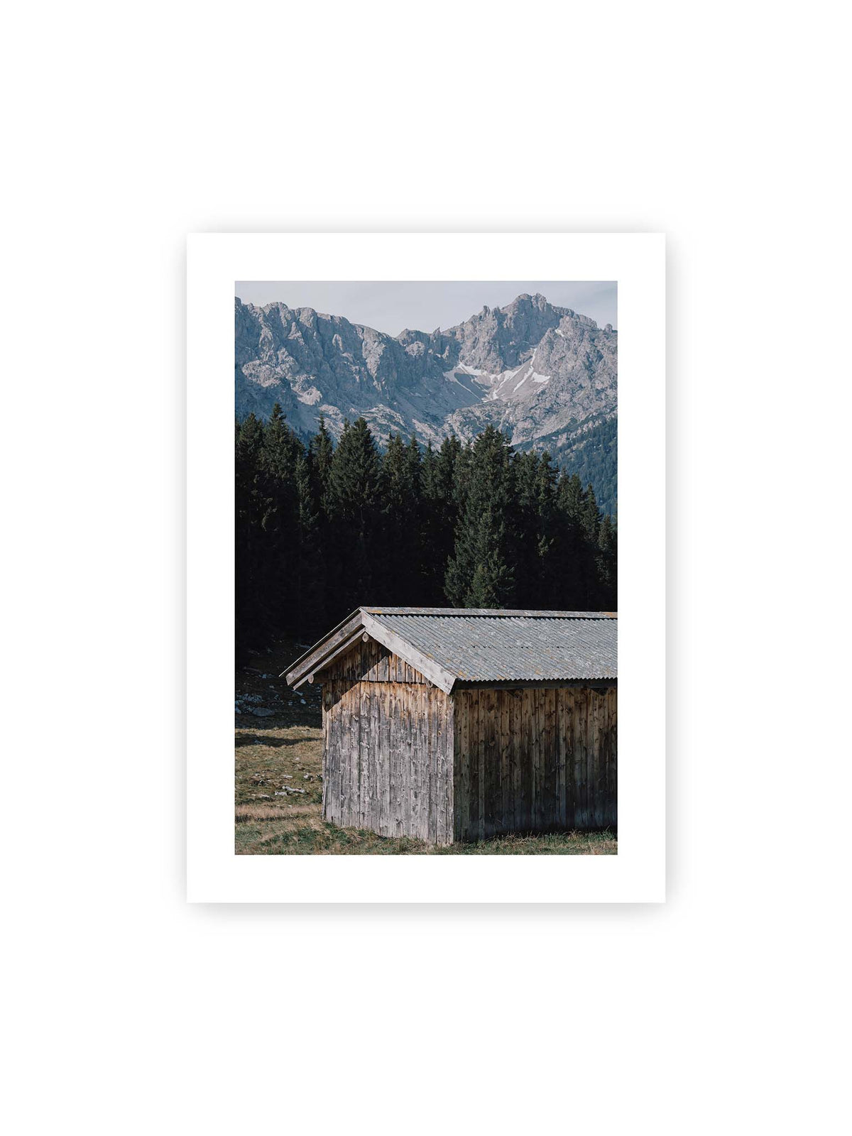 50x70 cm, weißer Galerie-Rand – Verwitterte Holzhütte vor dunklen Nadelbäumen und einer hellen, zerklüfteten Bergkulisse.