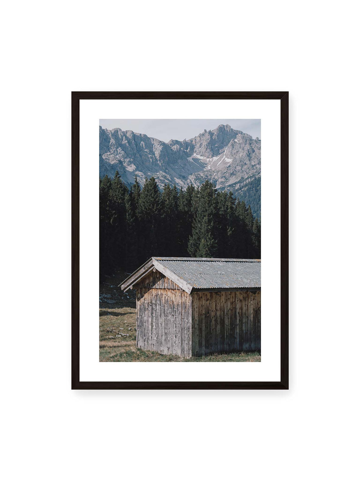 50x70 cm, weißer Galerie-Rand, schwarzer Rahmen – Verwitterte Holzhütte vor dunklen Nadelbäumen und einer hellen, zerklüfteten Bergkulisse.