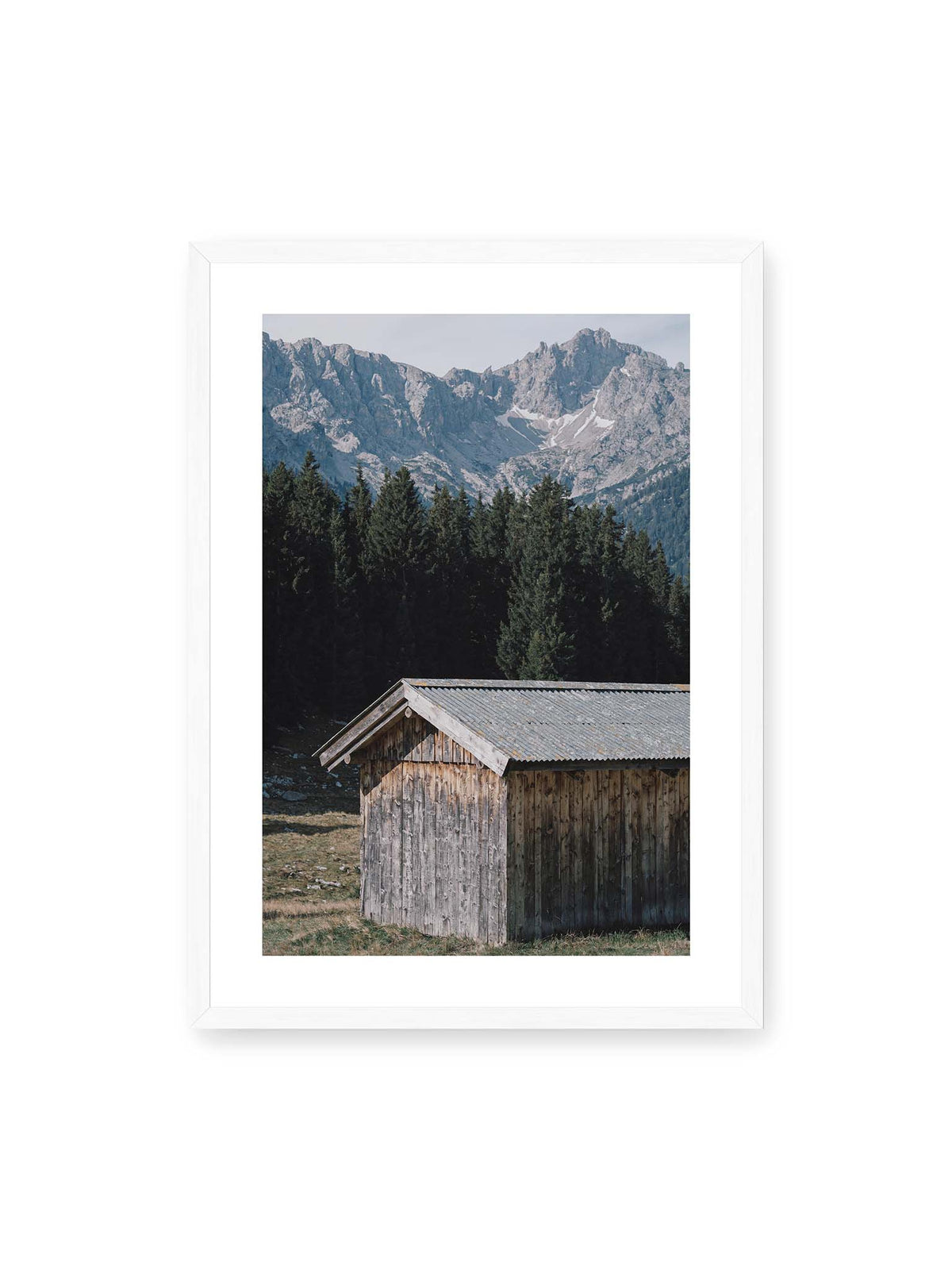 50x70 cm, weißer Galerie-Rand, weißer Rahmen – Verwitterte Holzhütte vor dunklen Nadelbäumen und einer hellen, zerklüfteten Bergkulisse.
