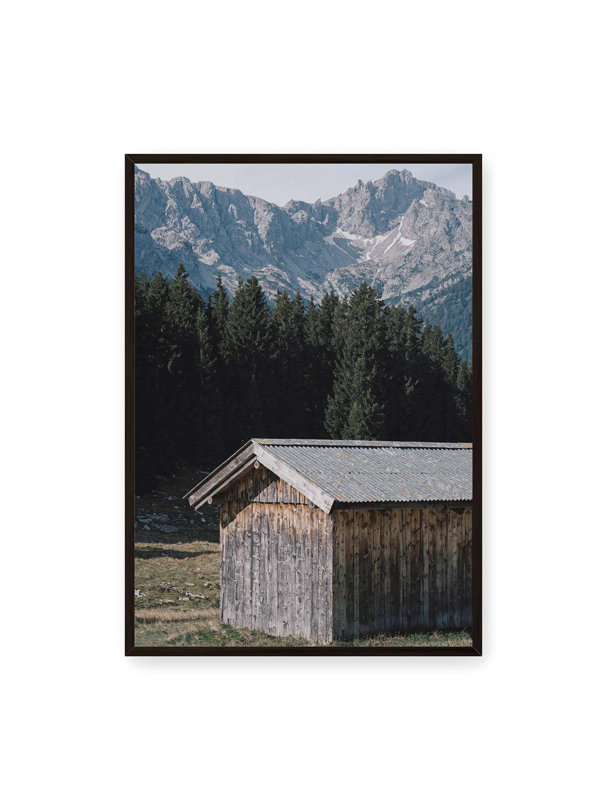 70x100 cm, schwarzer Rahmen – Verwitterte Holzhütte vor dunklen Nadelbäumen und einer hellen, zerklüfteten Bergkulisse.