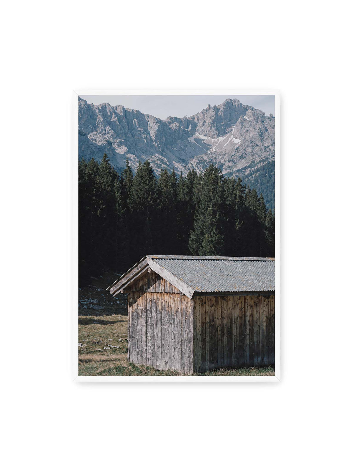 70x100 cm, weißer Rahmen – Verwitterte Holzhütte vor dunklen Nadelbäumen und einer hellen, zerklüfteten Bergkulisse.