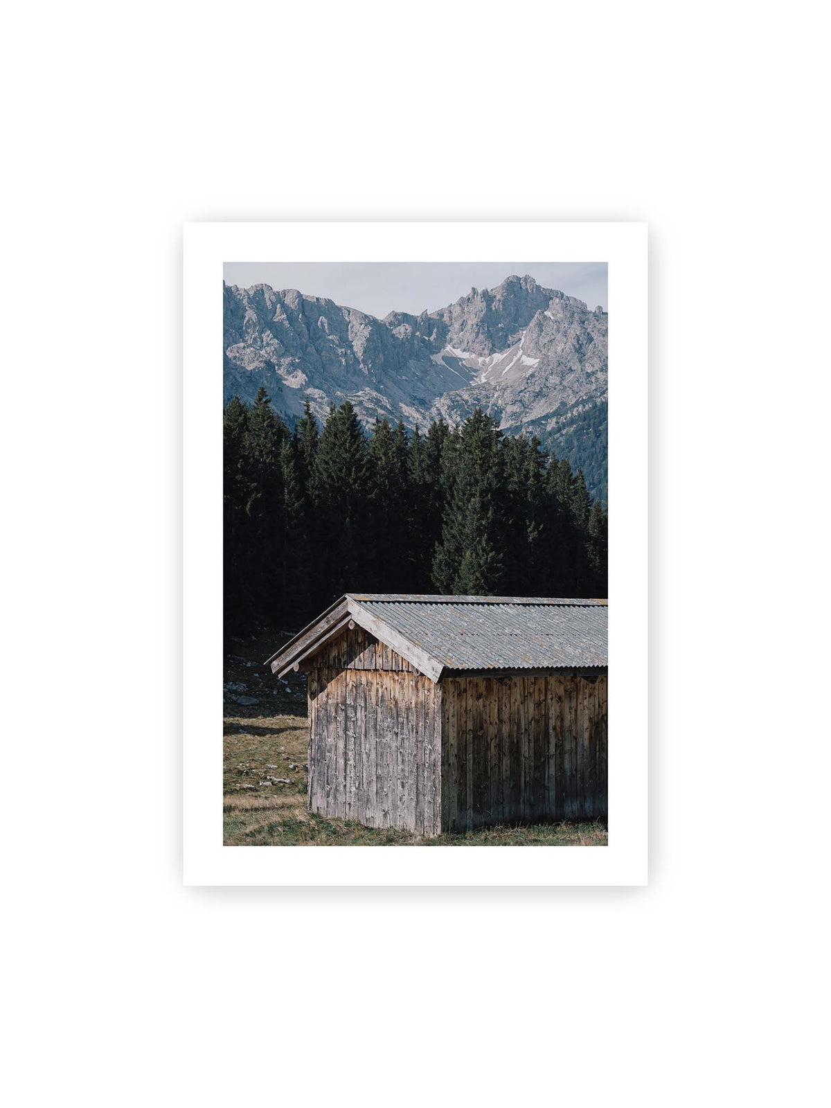 70x100 cm, weißer Galerie-Rand – Verwitterte Holzhütte vor dunklen Nadelbäumen und einer hellen, zerklüfteten Bergkulisse.