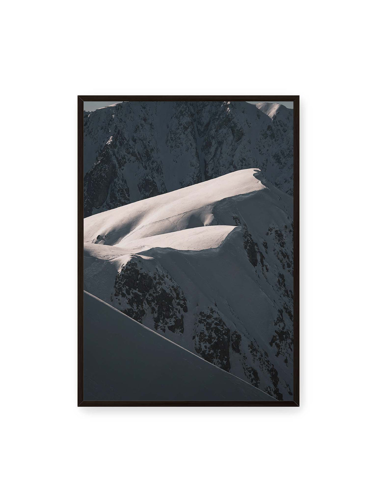 70x100 cm, schwarzer Rahmen – Verschneite Bergrücken mit hellen Graten und tiefen Schatten vor einer dunklen, steilen Felskulisse.