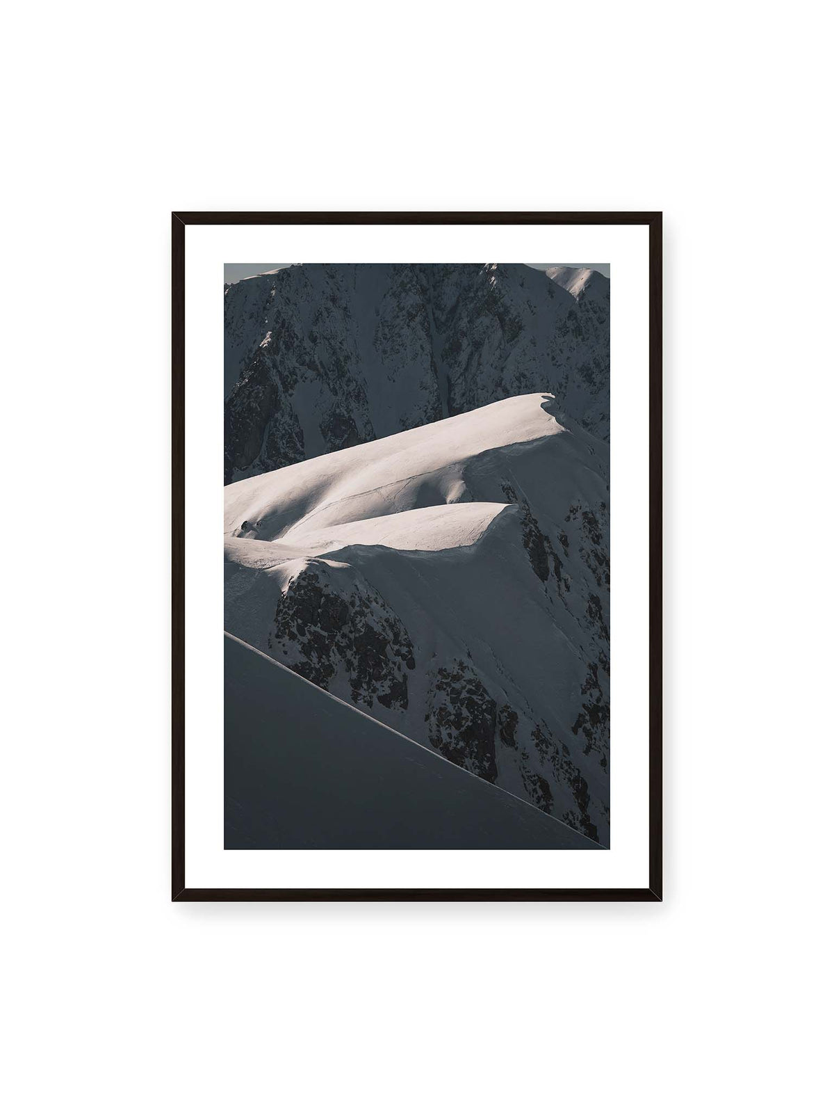 70x100 cm, weißer Galerie-Rand, schwarzer Rahmen – Verschneite Bergrücken mit hellen Graten und tiefen Schatten vor einer dunklen, steilen Felskulisse.