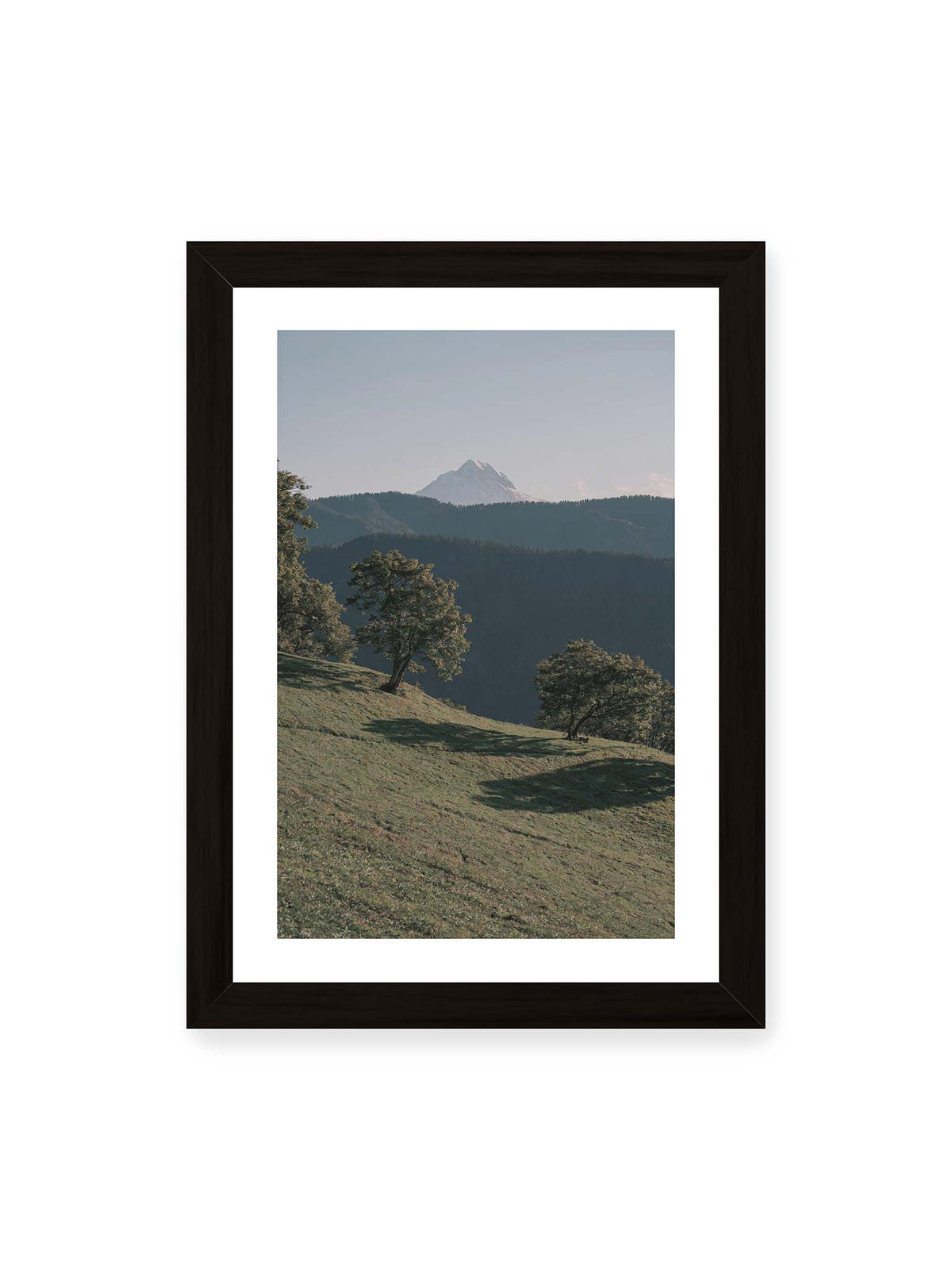 21x30 cm, weißer Galerie-Rand, schwarzer Rahmen – Ferner heller Berggipfel über dunklen Höhenzügen und einer geneigten grünen Wiese mit einzelnen Bäumen.