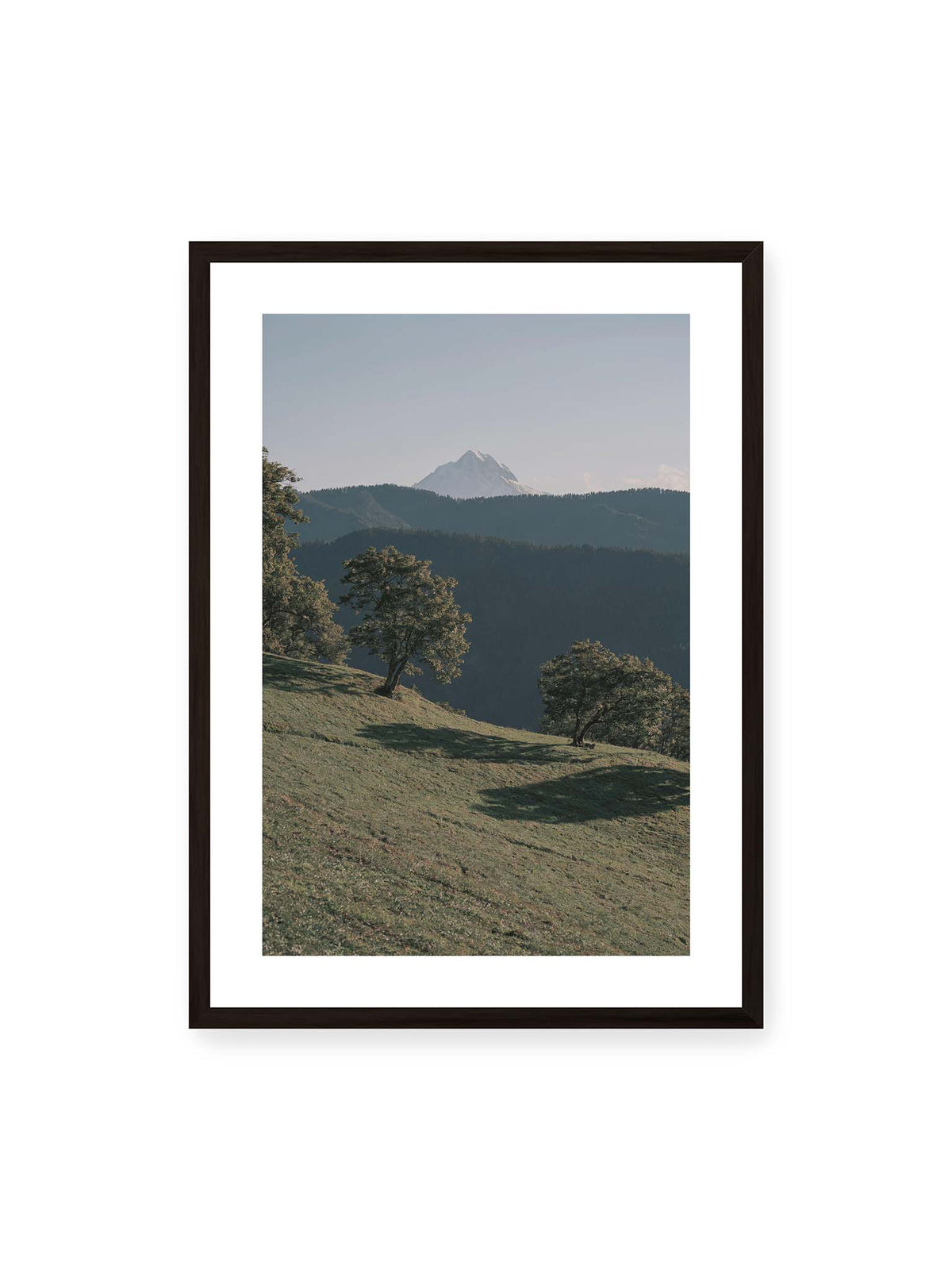 50x70 cm, weißer Galerie-Rand, schwarzer Rahmen – Ferner heller Berggipfel über dunklen Höhenzügen und einer geneigten grünen Wiese mit einzelnen Bäumen.