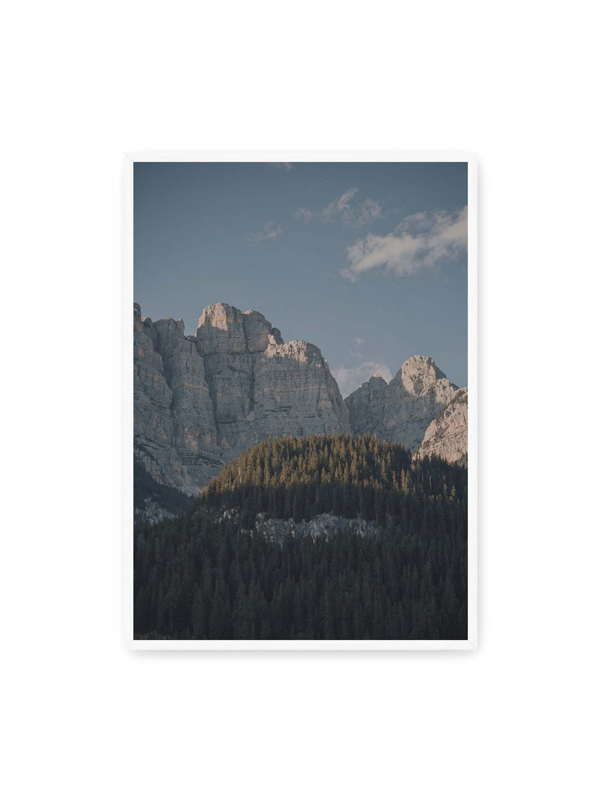 70x100 cm, weißer Rahmen – Helle Felswände erheben sich über einem dunklen Nadelwald vor klarem blauem Himmel.