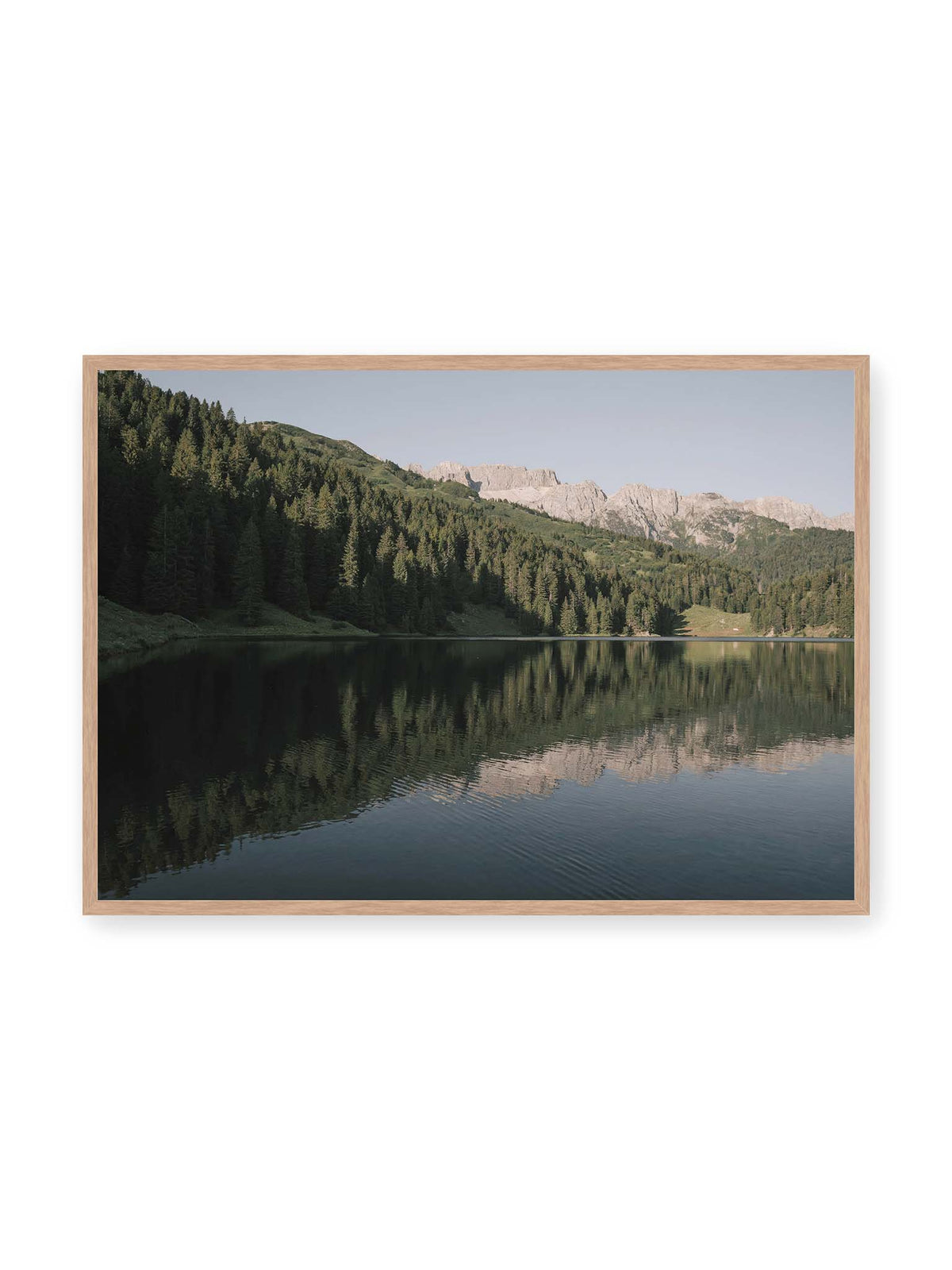 100x70 cm, Eichenrahmen – Ruhiger Bergsee mit Spiegelungen von bewaldeten Hängen und einer hellen Felskette unter blauem Himmel.