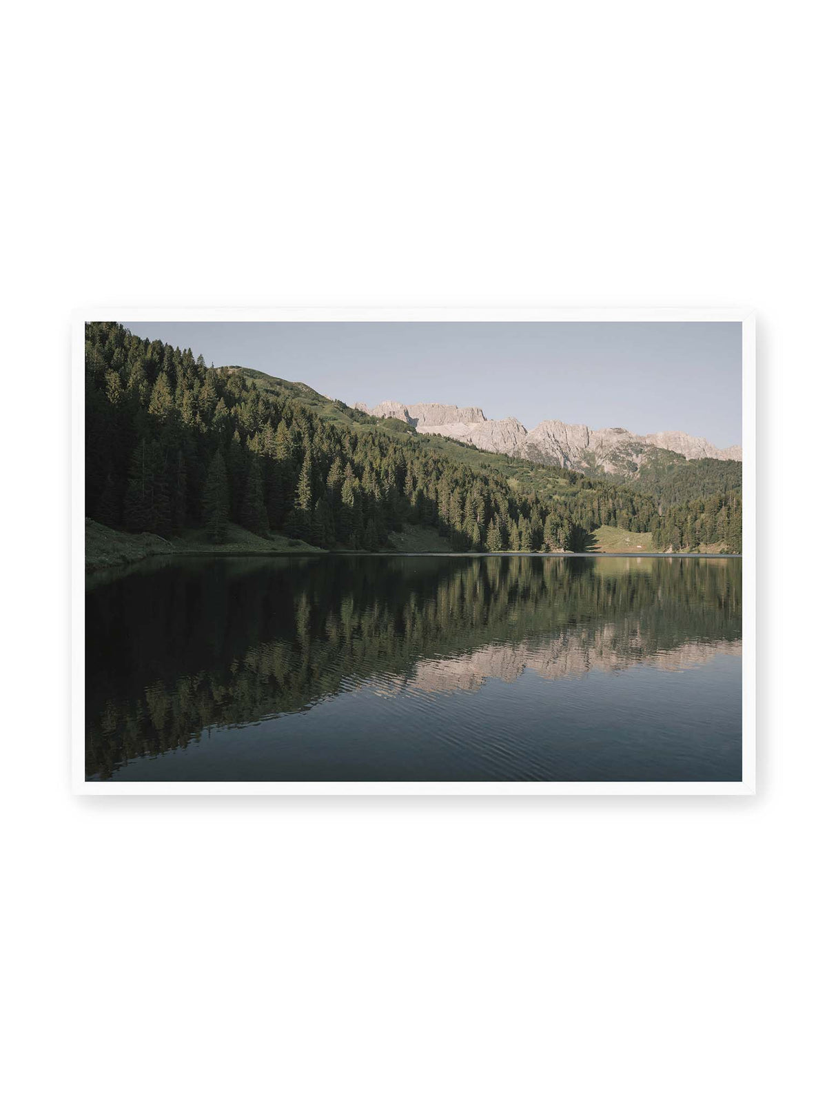 100x70 cm, weißer Rahmen – Ruhiger Bergsee mit Spiegelungen von bewaldeten Hängen und einer hellen Felskette unter blauem Himmel.