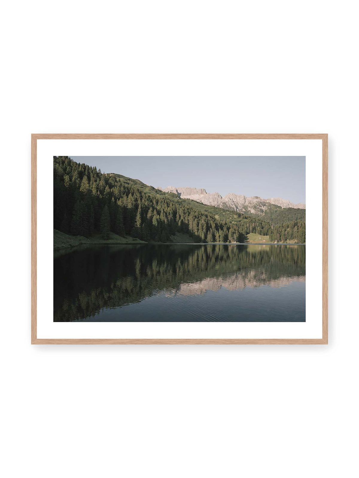 100x70 cm, weißer Galerie-Rand, Eichenrahmen – Ruhiger Bergsee mit Spiegelungen von bewaldeten Hängen und einer hellen Felskette unter blauem Himmel.