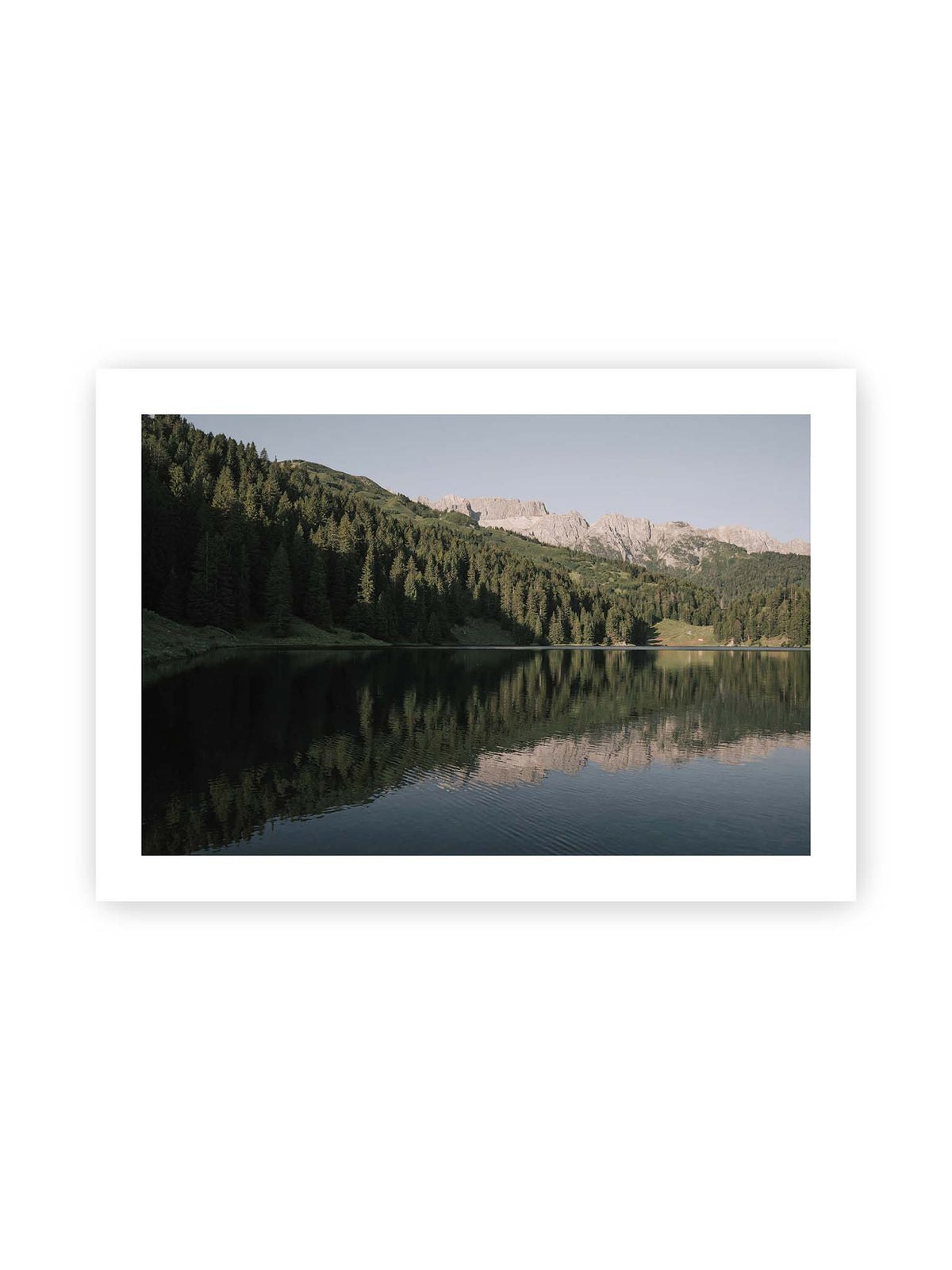 100x70 cm, weißer Galerie-Rand – Ruhiger Bergsee mit Spiegelungen von bewaldeten Hängen und einer hellen Felskette unter blauem Himmel.
