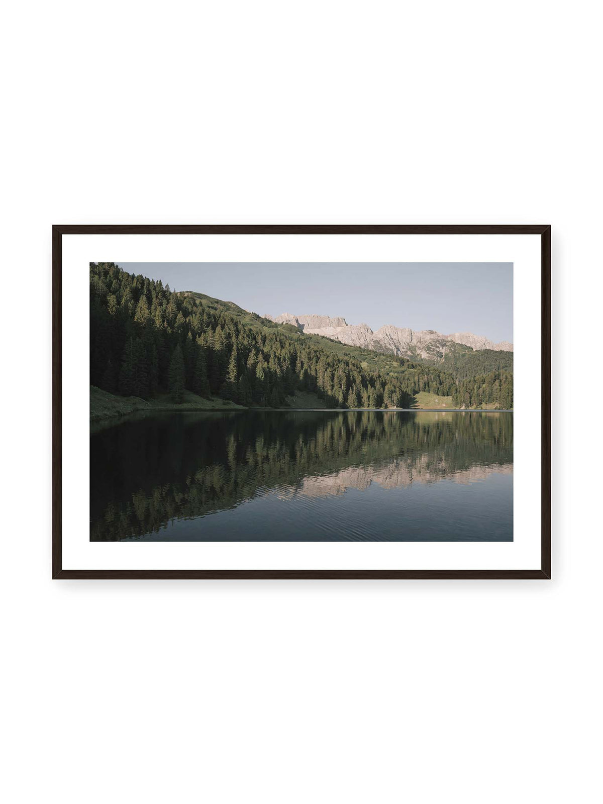 100x70 cm, weißer Galerie-Rand, schwarzer Rahmen – Ruhiger Bergsee mit Spiegelungen von bewaldeten Hängen und einer hellen Felskette unter blauem Himmel.