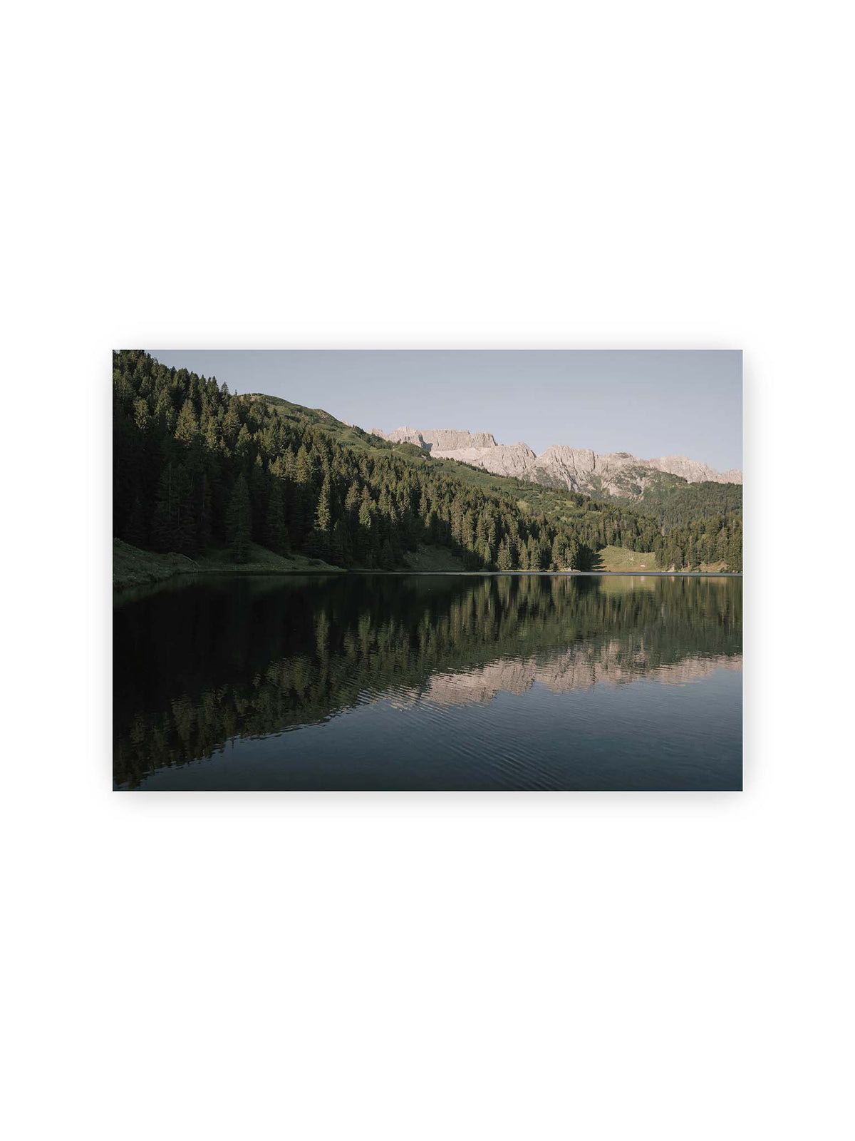 30x21 cm – Ruhiger Bergsee mit Spiegelungen von bewaldeten Hängen und einer hellen Felskette unter blauem Himmel.