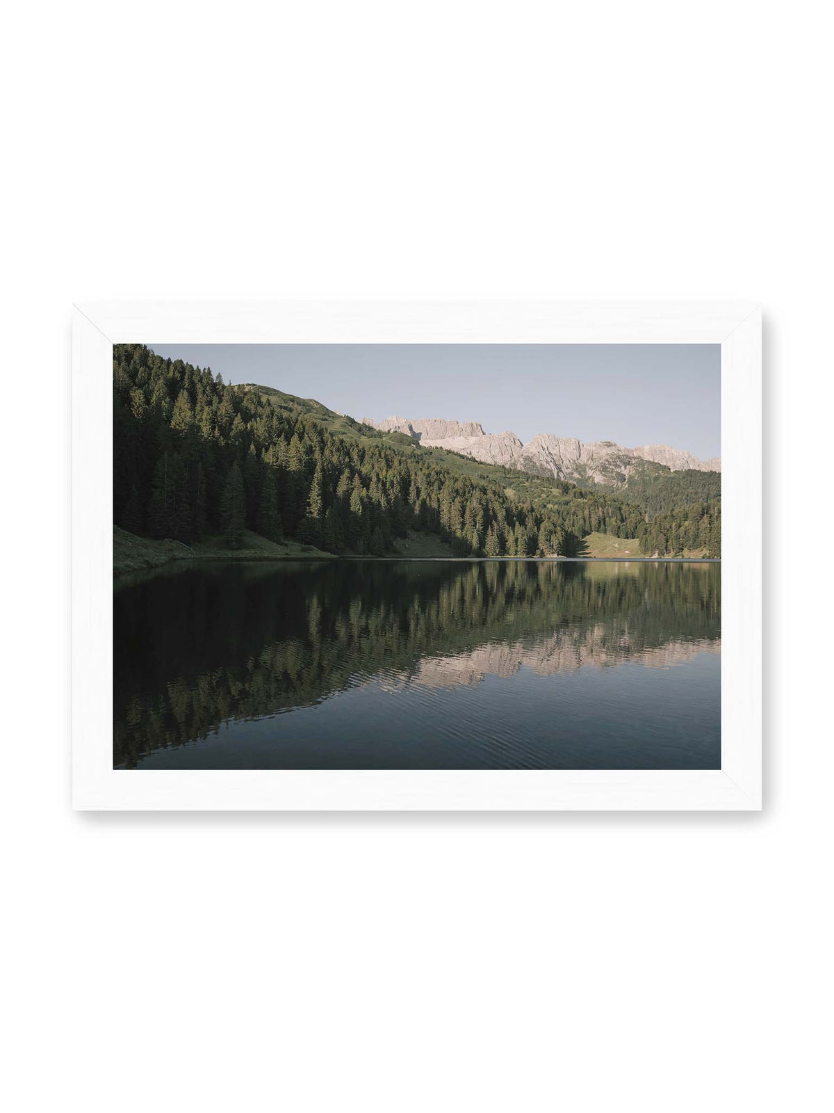 30x21 cm, weißer Rahmen – Ruhiger Bergsee mit Spiegelungen von bewaldeten Hängen und einer hellen Felskette unter blauem Himmel.