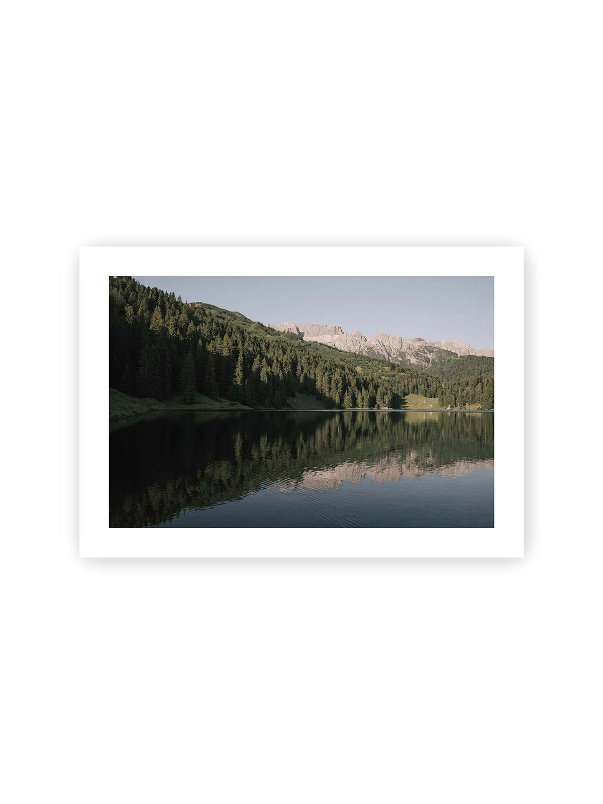 30x21 cm, weißer Galerie-Rand – Ruhiger Bergsee mit Spiegelungen von bewaldeten Hängen und einer hellen Felskette unter blauem Himmel.