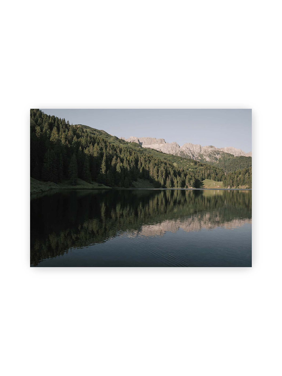 70x50 cm – Ruhiger Bergsee mit Spiegelungen von bewaldeten Hängen und einer hellen Felskette unter blauem Himmel.