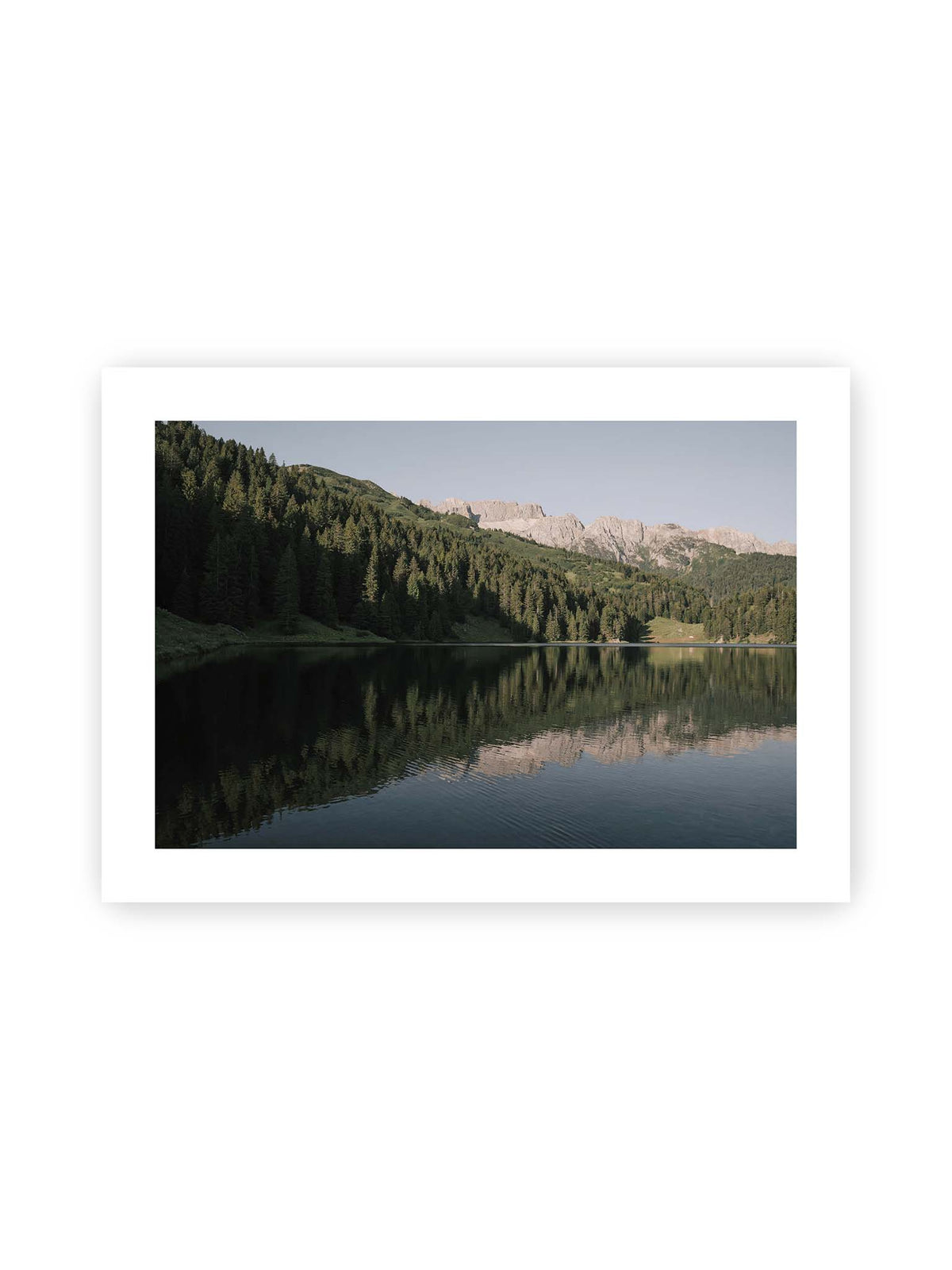 70x50 cm, weißer Galerie-Rand – Ruhiger Bergsee mit Spiegelungen von bewaldeten Hängen und einer hellen Felskette unter blauem Himmel.