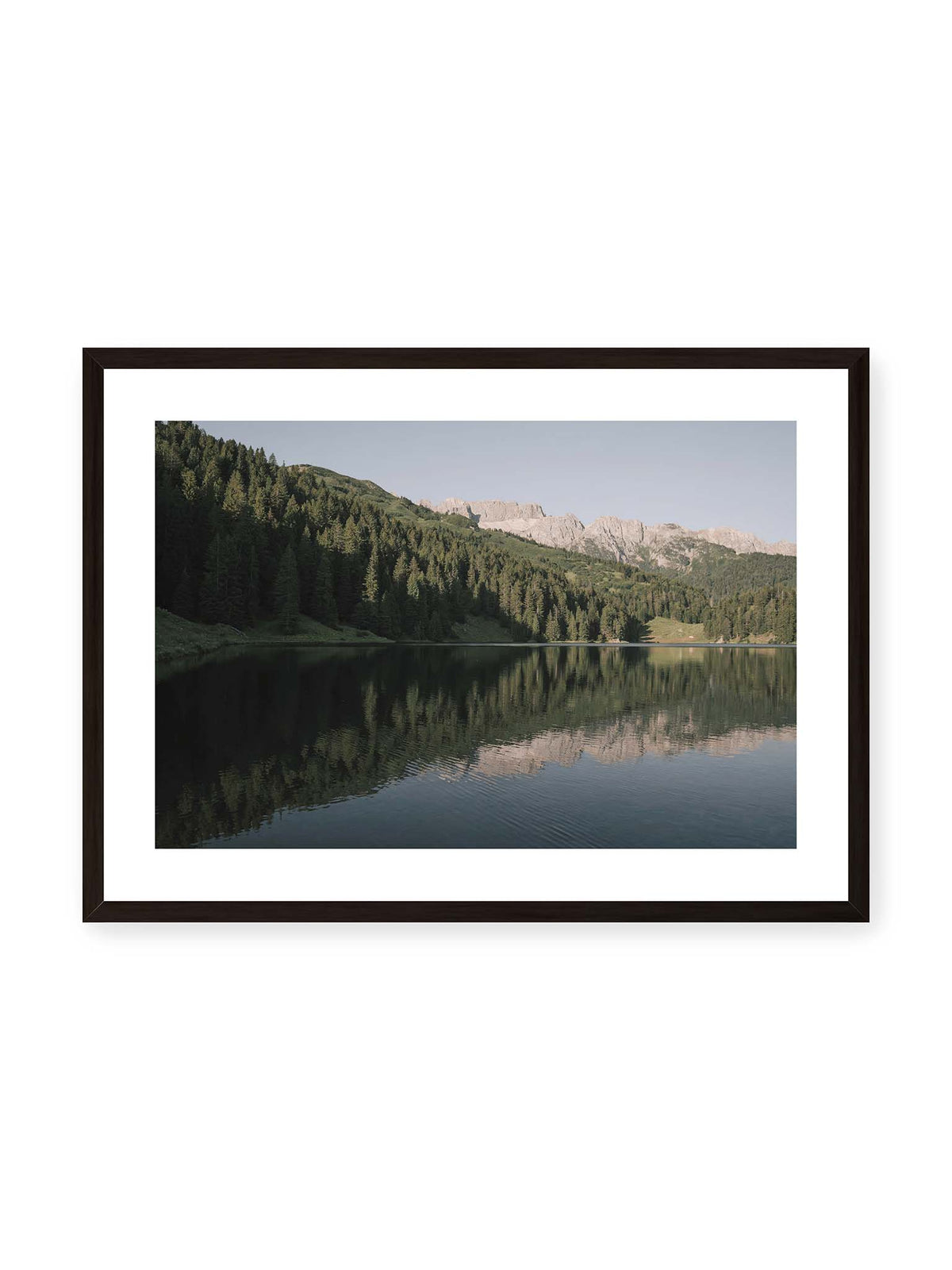 70x50 cm, weißer Galerie-Rand, schwarzer Rahmen – Ruhiger Bergsee mit Spiegelungen von bewaldeten Hängen und einer hellen Felskette unter blauem Himmel.