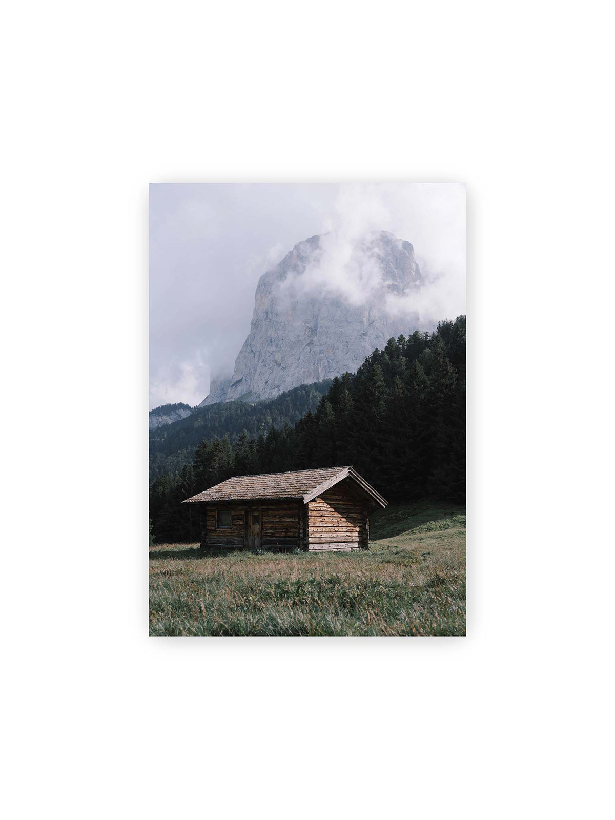 21x30 cm – Holzhütte auf einer Wiese vor dichtem Nadelwald und einer hohen, teilweise von Wolken verhüllten Felswand.