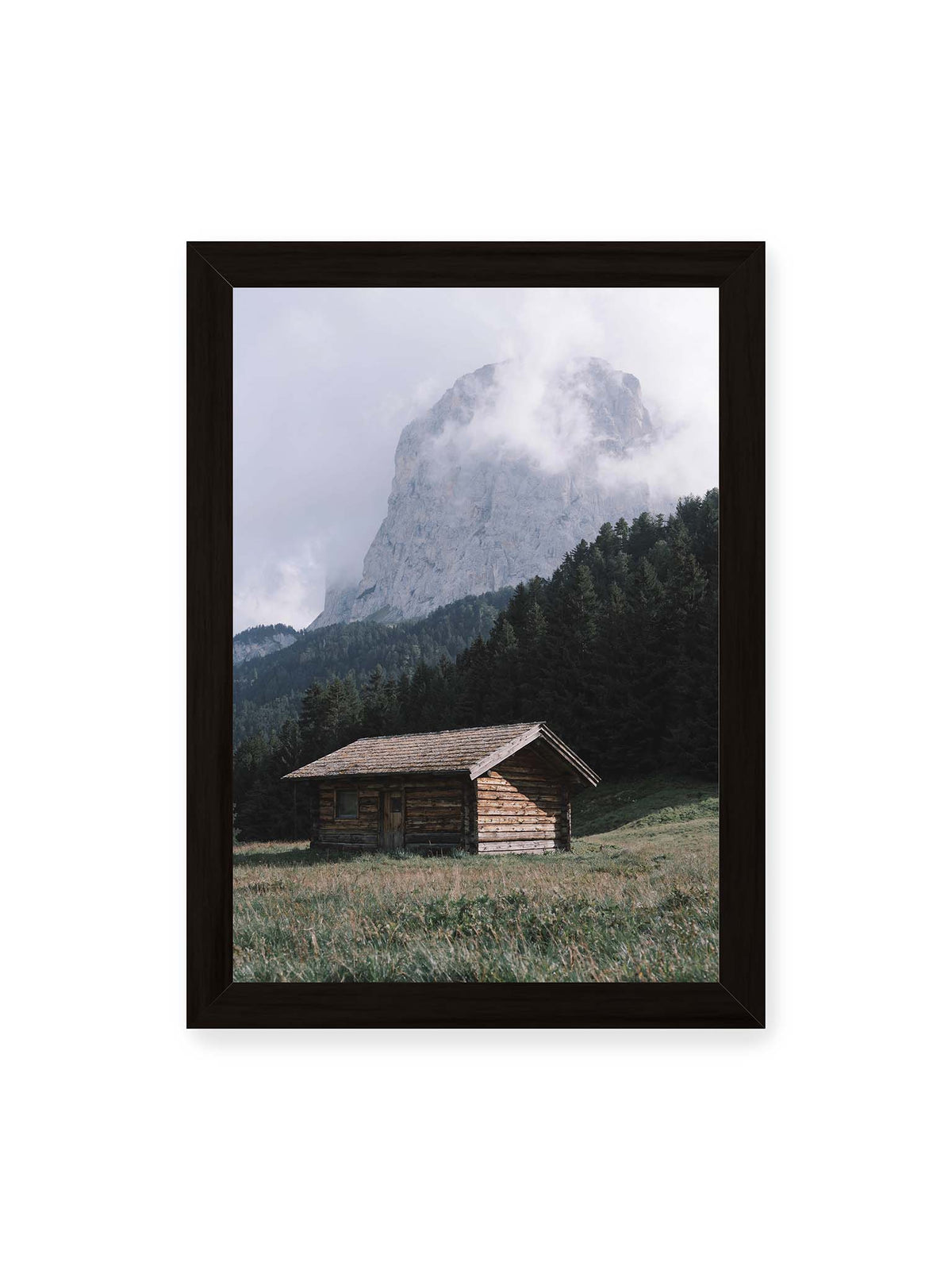 21x30 cm, schwarzer Rahmen – Holzhütte auf einer Wiese vor dichtem Nadelwald und einer hohen, teilweise von Wolken verhüllten Felswand.