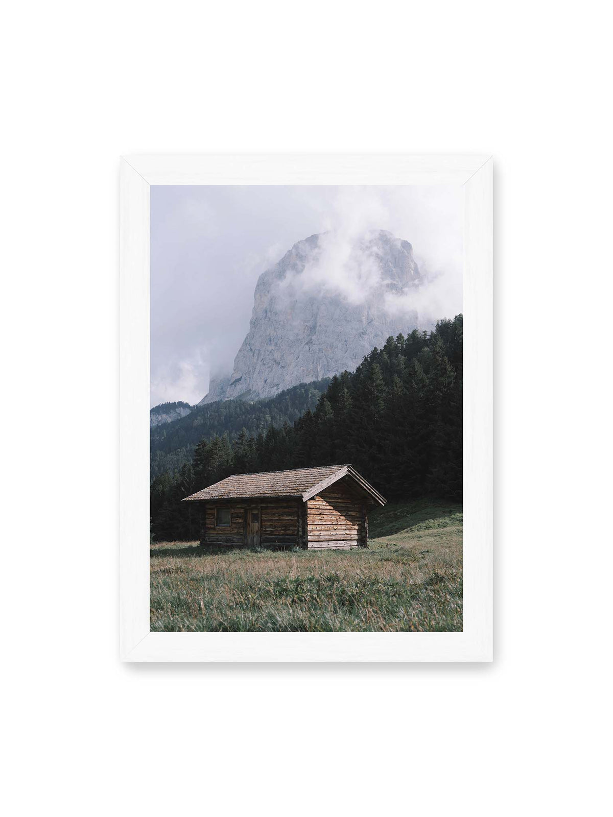 21x30 cm, weißer Rahmen – Holzhütte auf einer Wiese vor dichtem Nadelwald und einer hohen, teilweise von Wolken verhüllten Felswand.