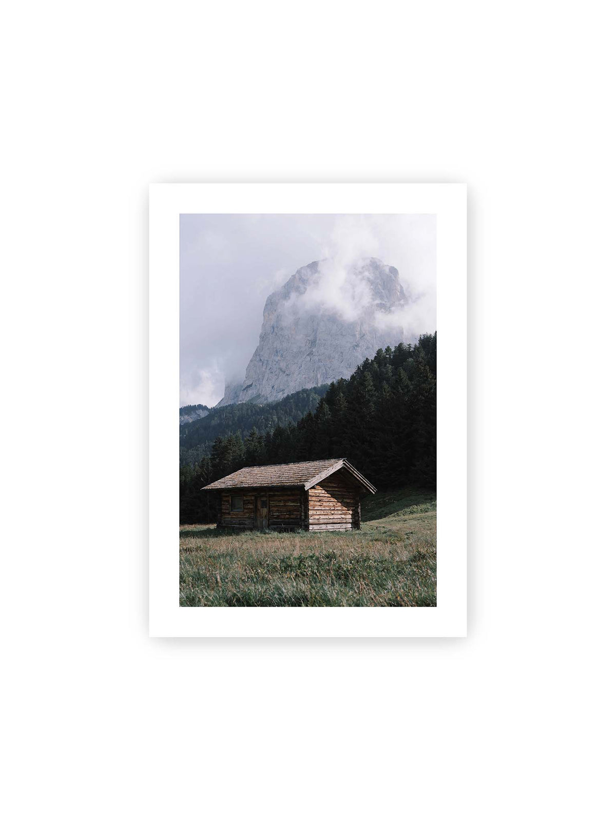 21x30 cm, weißer Galerie-Rand – Holzhütte auf einer Wiese vor dichtem Nadelwald und einer hohen, teilweise von Wolken verhüllten Felswand.
