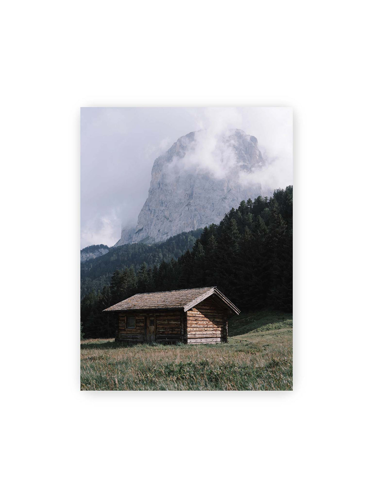30x40 cm – Holzhütte auf einer Wiese vor dichtem Nadelwald und einer hohen, teilweise von Wolken verhüllten Felswand.