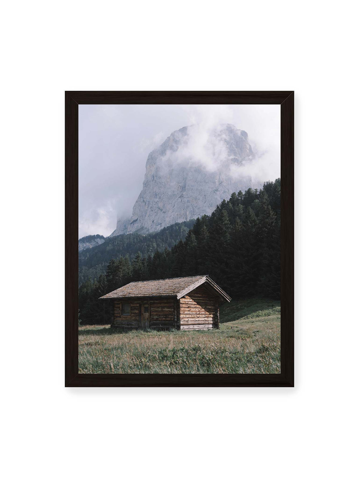 30x40 cm, schwarzer Rahmen – Holzhütte auf einer Wiese vor dichtem Nadelwald und einer hohen, teilweise von Wolken verhüllten Felswand.
