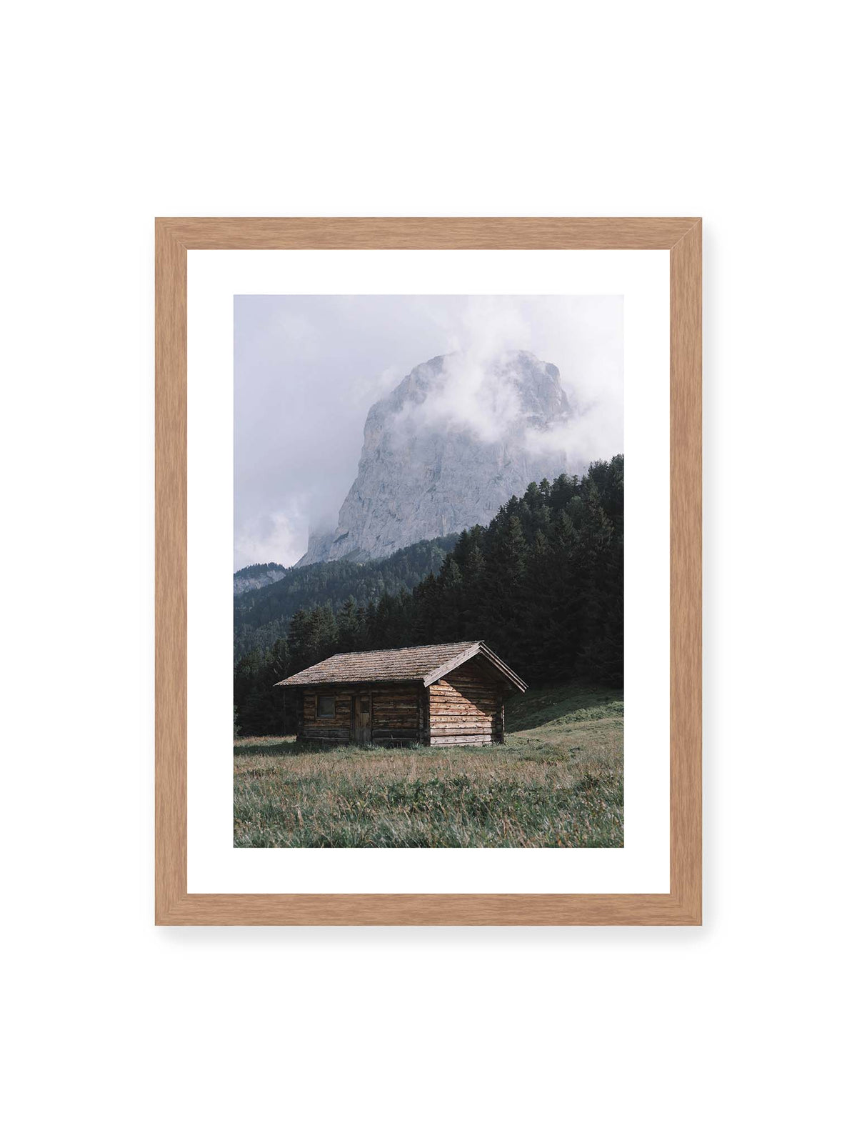 30x40 cm, weißer Galerie-Rand, Eichenrahmen – Holzhütte auf einer Wiese vor dichtem Nadelwald und einer hohen, teilweise von Wolken verhüllten Felswand.