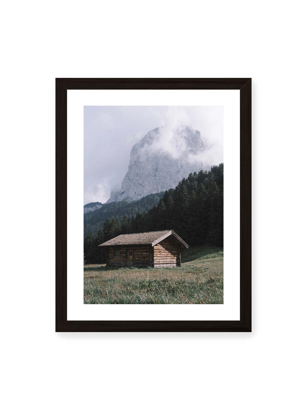 30x40 cm, weißer Galerie-Rand, schwarzer Rahmen – Holzhütte auf einer Wiese vor dichtem Nadelwald und einer hohen, teilweise von Wolken verhüllten Felswand.