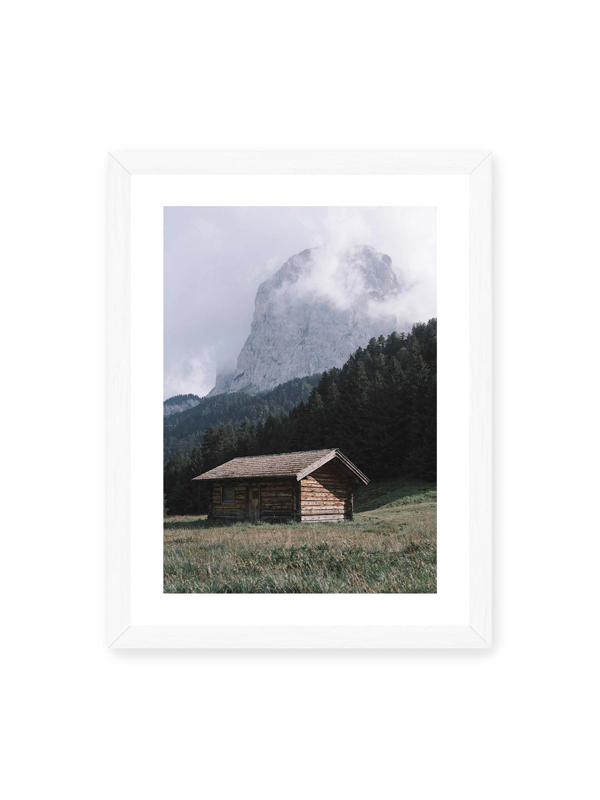 30x40 cm, weißer Galerie-Rand, weißer Rahmen – Holzhütte auf einer Wiese vor dichtem Nadelwald und einer hohen, teilweise von Wolken verhüllten Felswand.