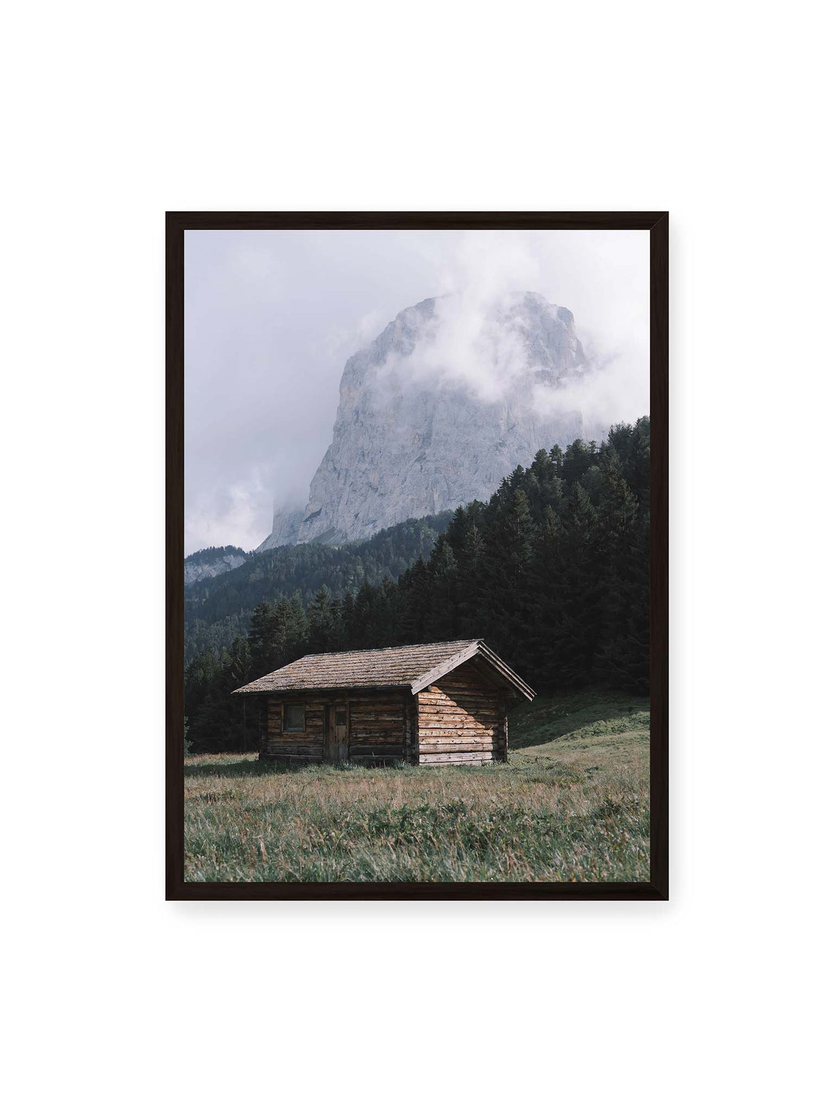 50x70 cm, schwarzer Rahmen – Holzhütte auf einer Wiese vor dichtem Nadelwald und einer hohen, teilweise von Wolken verhüllten Felswand.