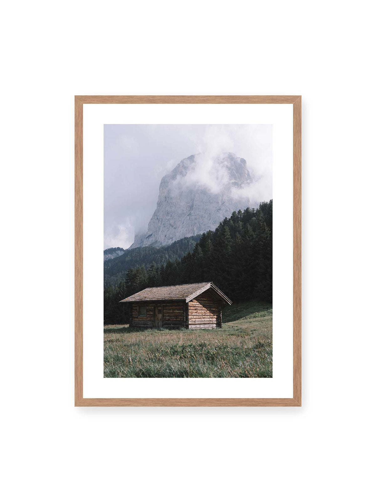 50x70 cm, weißer Galerie-Rand, Eichenrahmen – Holzhütte auf einer Wiese vor dichtem Nadelwald und einer hohen, teilweise von Wolken verhüllten Felswand.