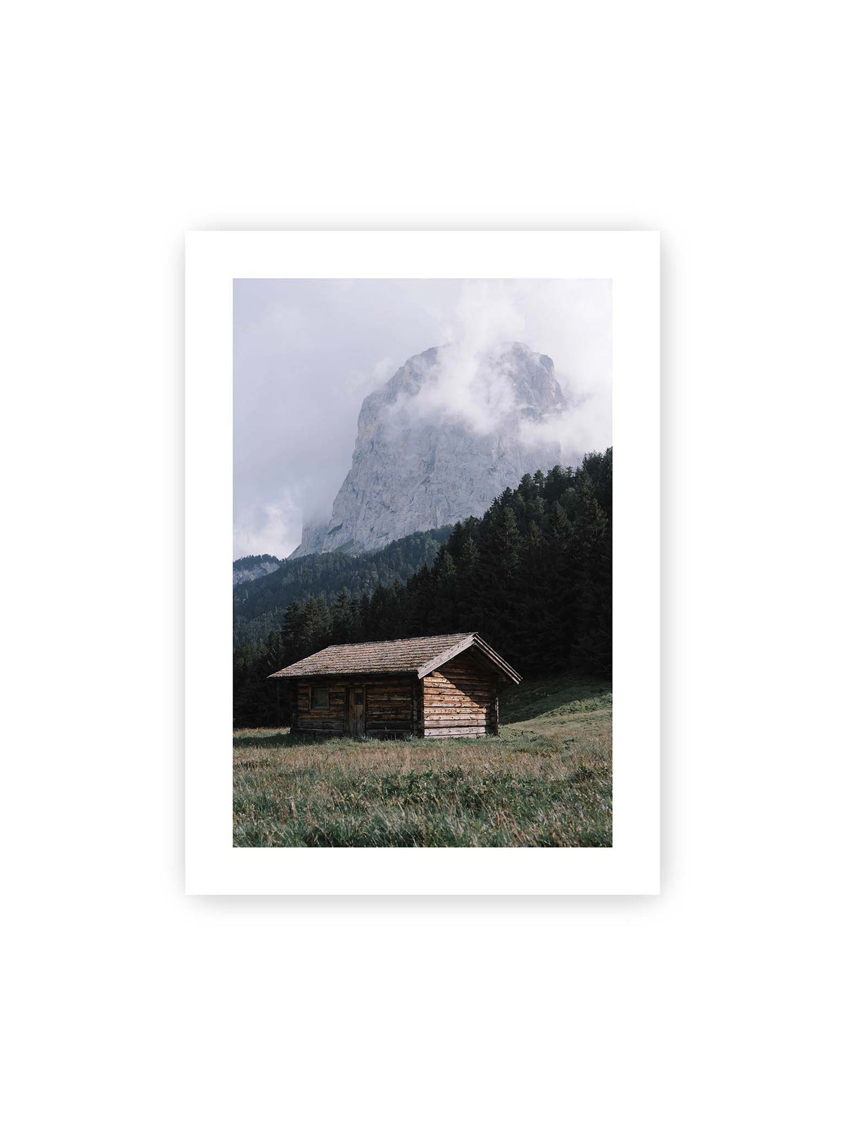50x70 cm, weißer Galerie-Rand – Holzhütte auf einer Wiese vor dichtem Nadelwald und einer hohen, teilweise von Wolken verhüllten Felswand.