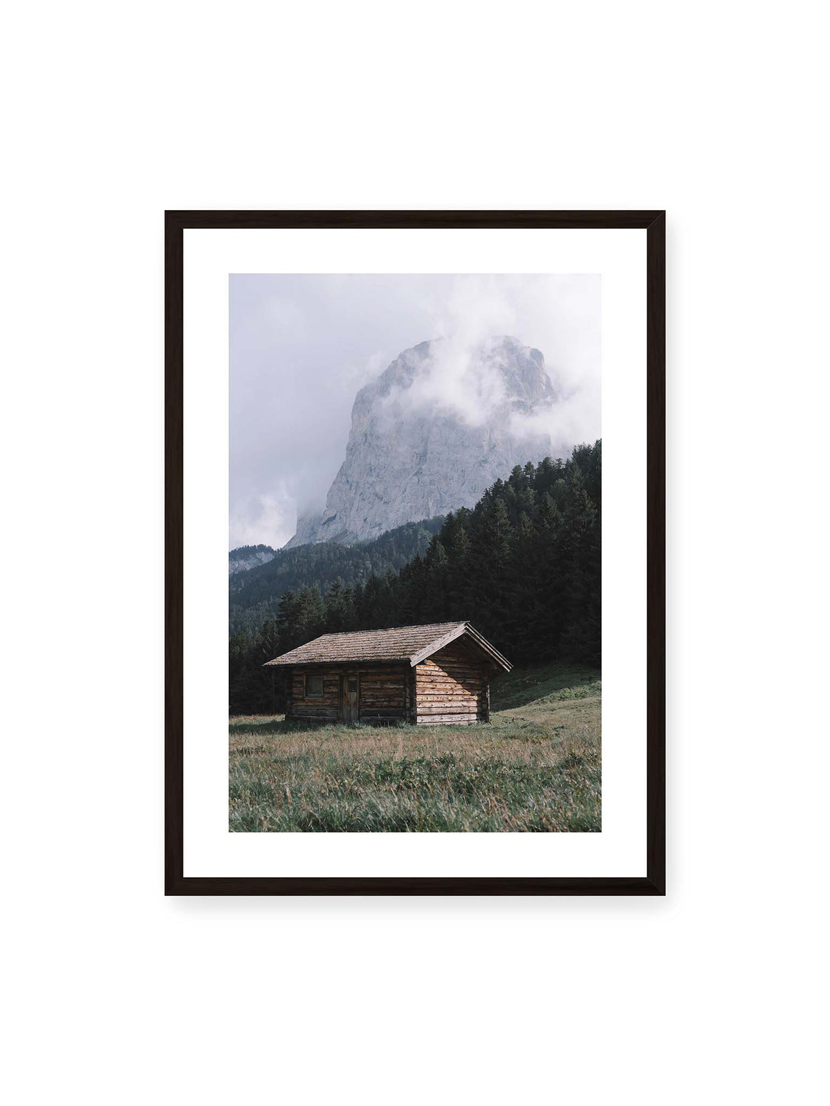 50x70 cm, weißer Galerie-Rand, schwarzer Rahmen – Holzhütte auf einer Wiese vor dichtem Nadelwald und einer hohen, teilweise von Wolken verhüllten Felswand.