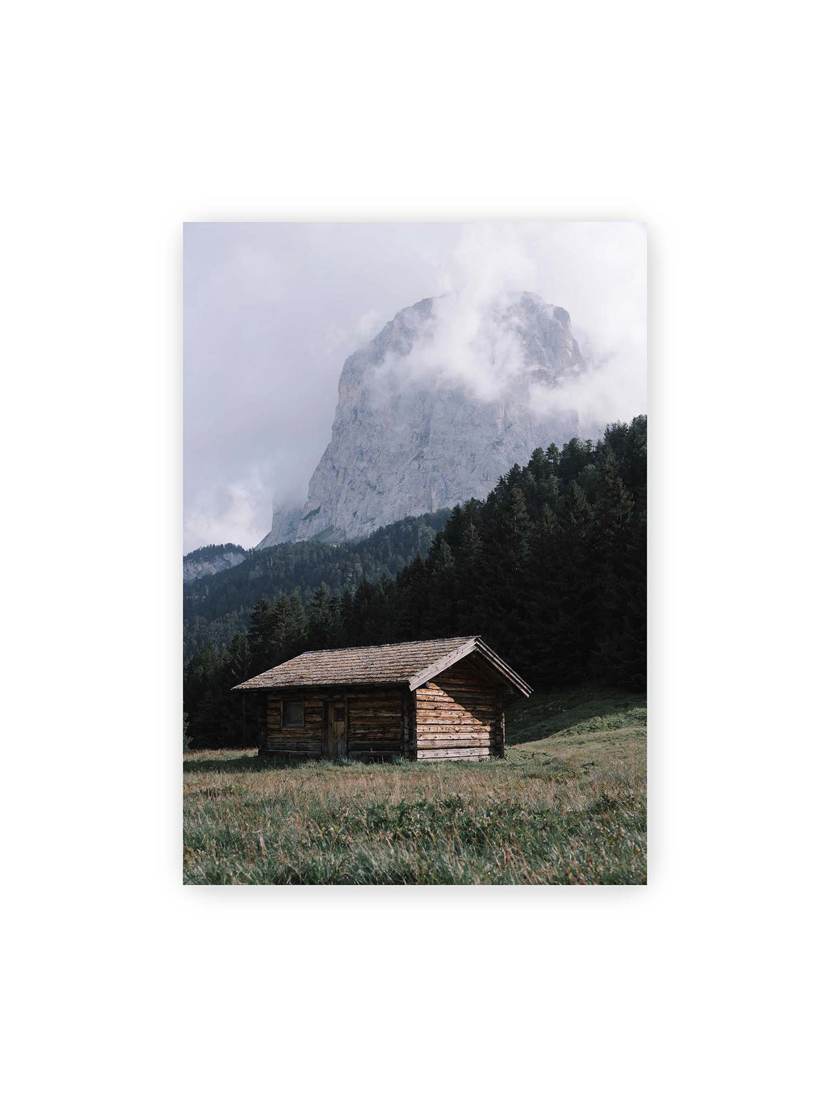 70x100 cm – Holzhütte auf einer Wiese vor dichtem Nadelwald und einer hohen, teilweise von Wolken verhüllten Felswand.