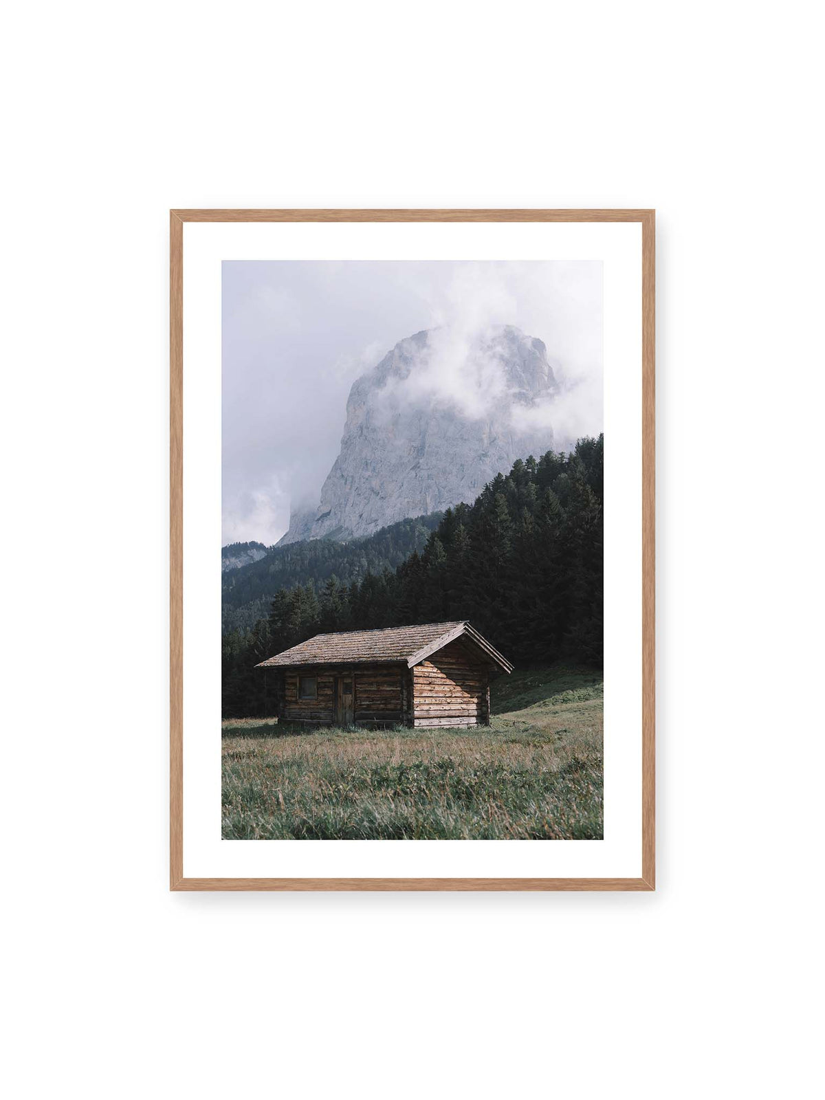 70x100 cm, weißer Galerie-Rand, Eichenrahmen – Holzhütte auf einer Wiese vor dichtem Nadelwald und einer hohen, teilweise von Wolken verhüllten Felswand.