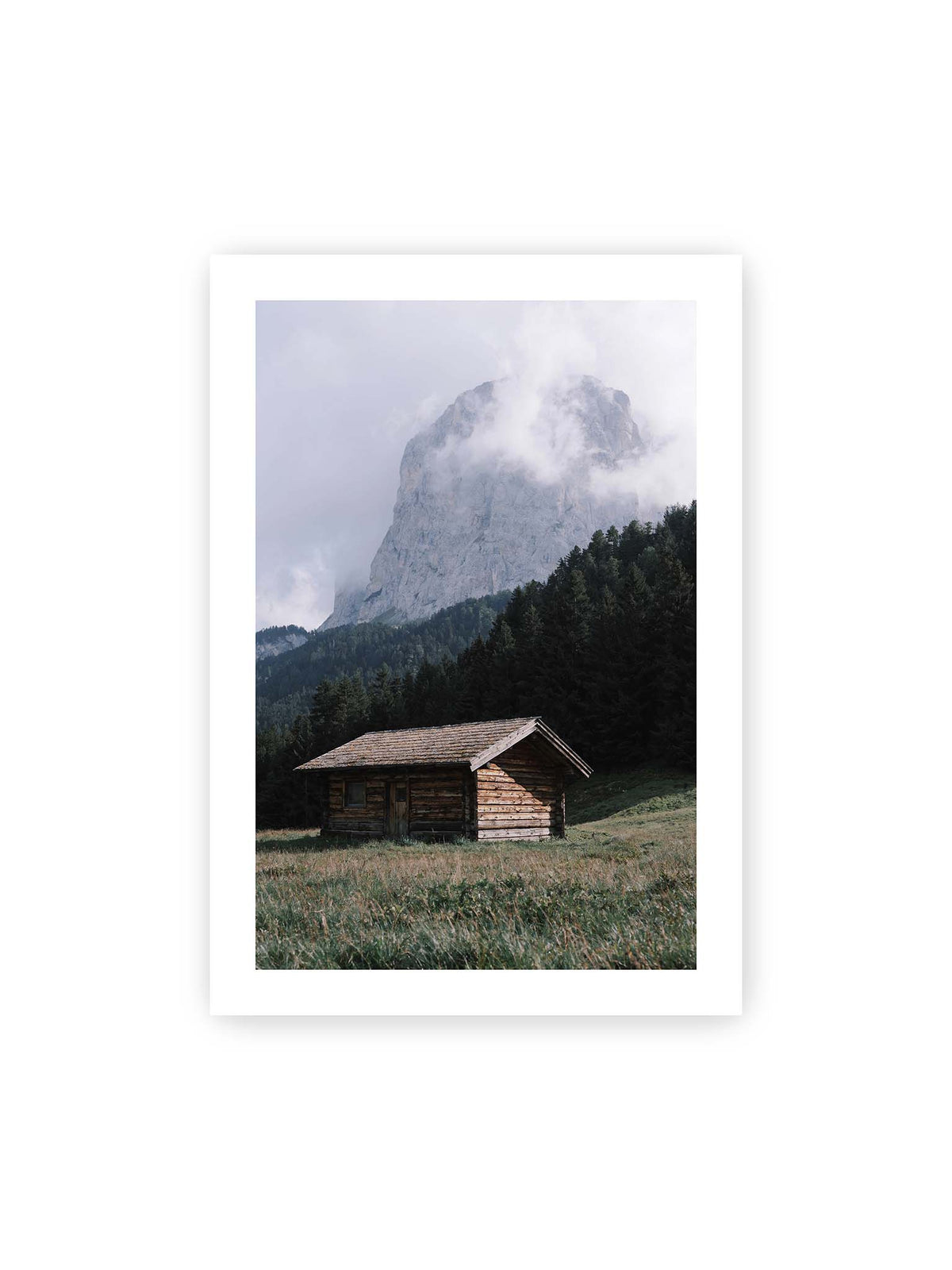 70x100 cm, weißer Galerie-Rand – Holzhütte auf einer Wiese vor dichtem Nadelwald und einer hohen, teilweise von Wolken verhüllten Felswand.