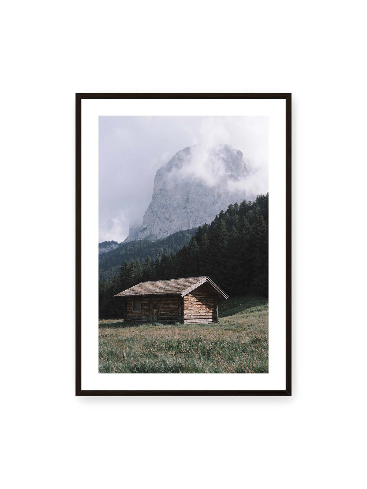 70x100 cm, weißer Galerie-Rand, schwarzer Rahmen – Holzhütte auf einer Wiese vor dichtem Nadelwald und einer hohen, teilweise von Wolken verhüllten Felswand.