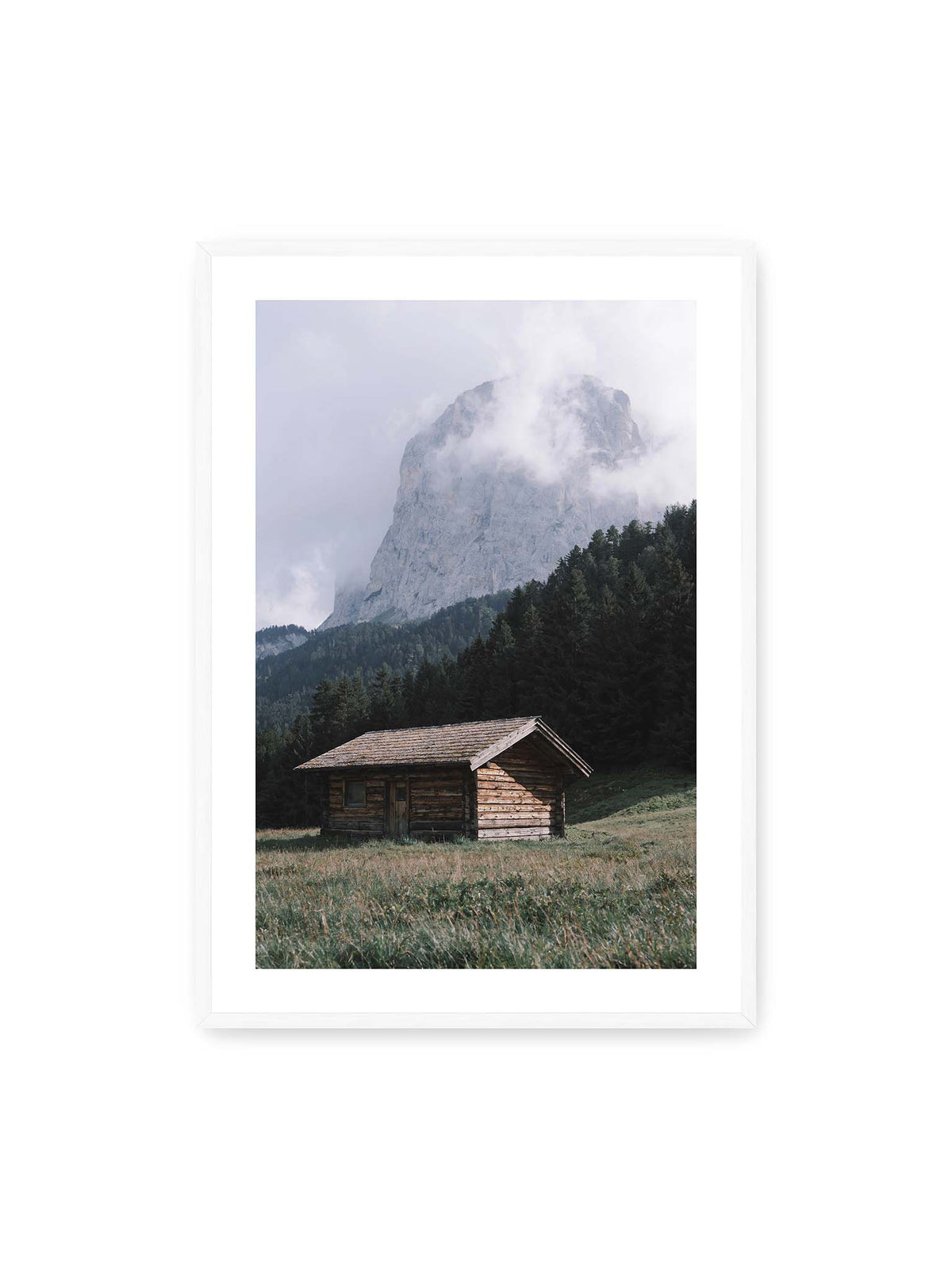 70x100 cm, weißer Galerie-Rand, weißer Rahmen – Holzhütte auf einer Wiese vor dichtem Nadelwald und einer hohen, teilweise von Wolken verhüllten Felswand.